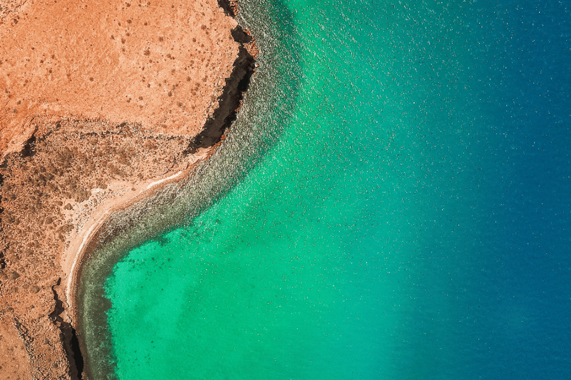 Vista aérea de una costa rocosa con una pequeña playa y aguas turquesas, donde el mar se encuentra con la tierra árida y rojiza.