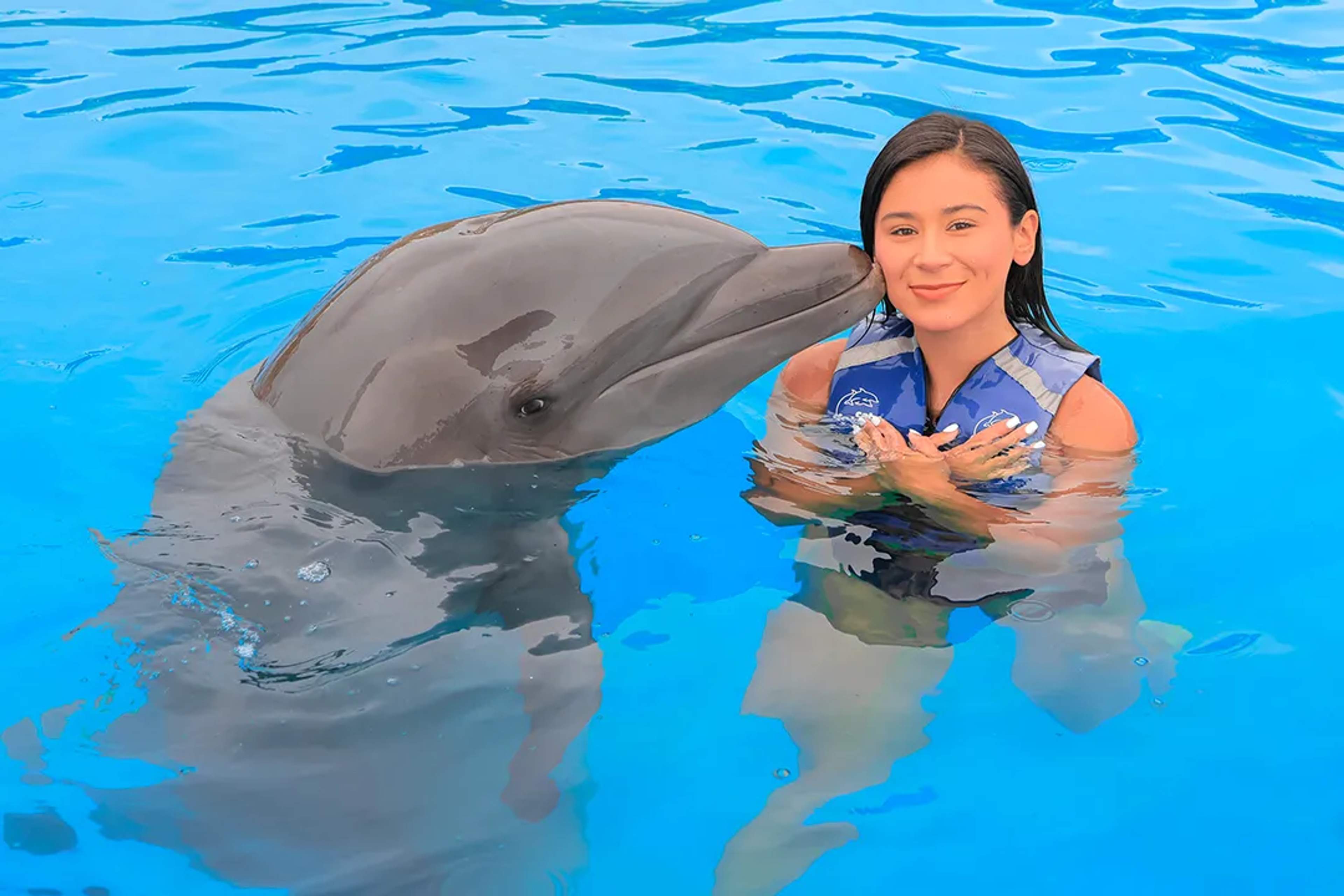 Woman in a life jacket smiles as a dolphin gives her a gentle kiss in a bright blue pool.