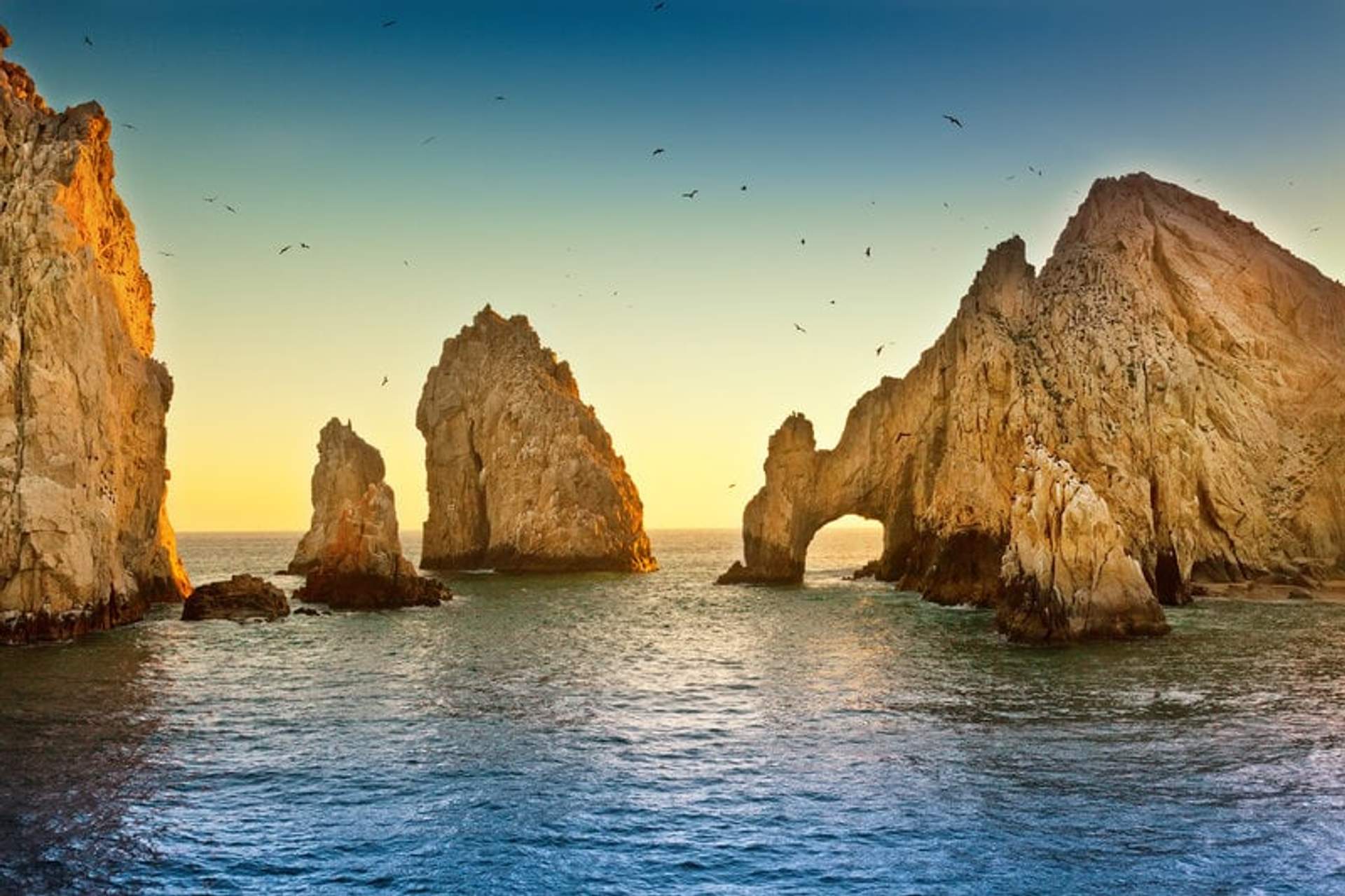 The iconic rock formations of Land's End in Cabo San Lucas bathed in the golden light of the setting sun, with birds soaring in the clear blue sky.