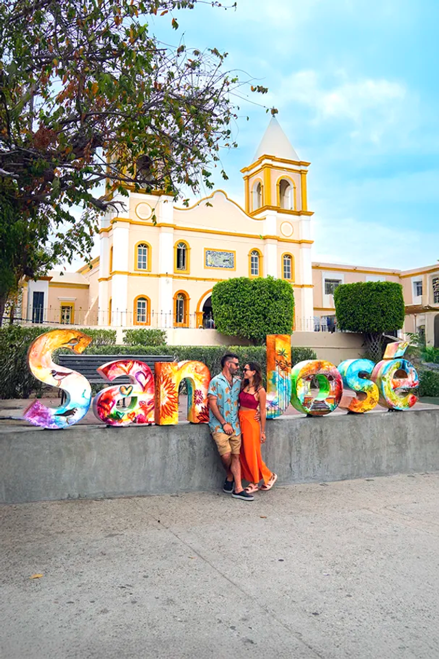 Tourists pose by a colorful "San José" sign in front of a historic church in Los Cabos.