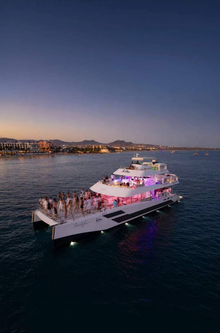Catamarán de lujo navegando al atardecer en la bahía de Cabo con invitados y luces de la ciudad.