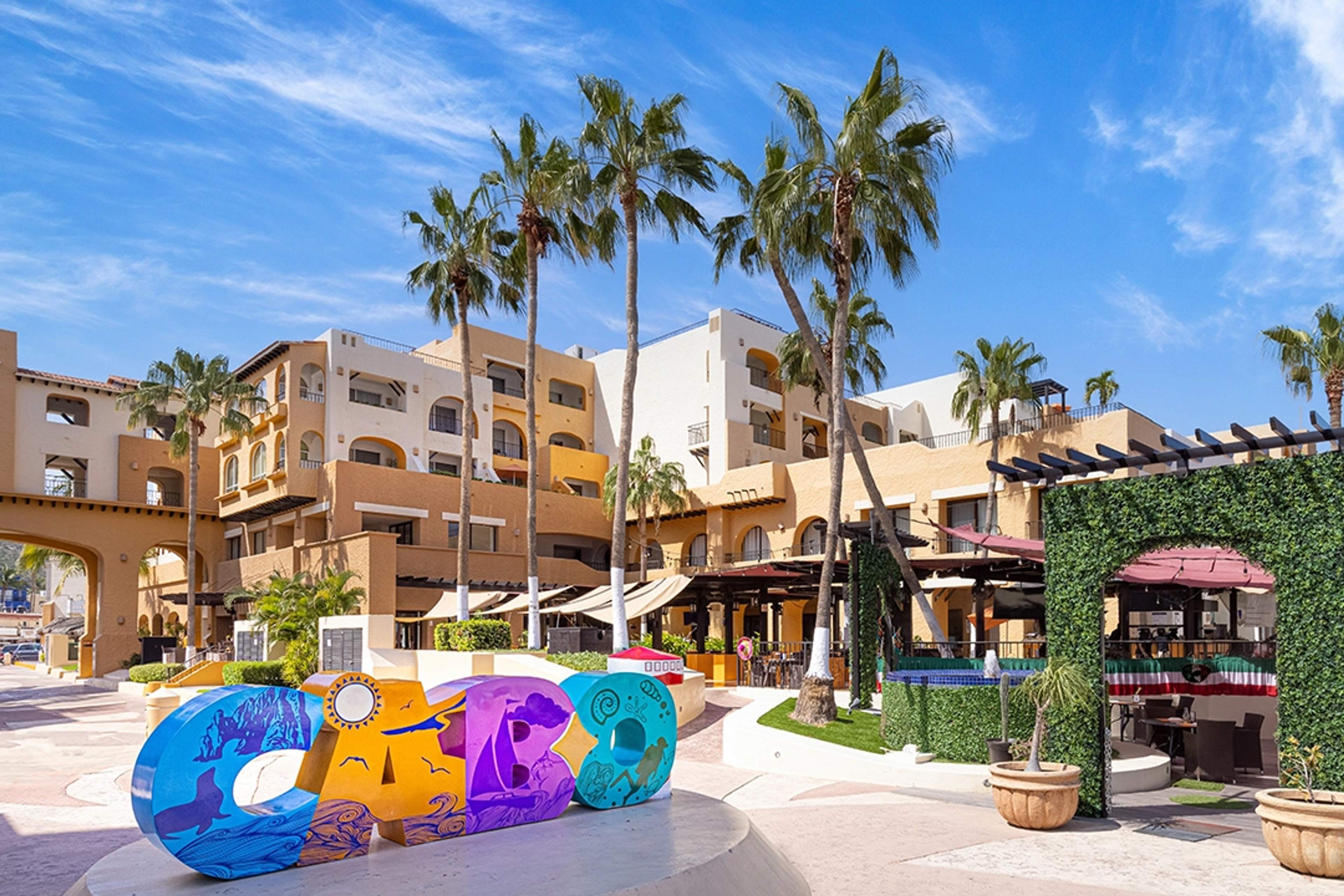 Colorful plaza in Cabo San Lucas featuring a vibrant "Cabo" sign, surrounded by palm trees and modern buildings.