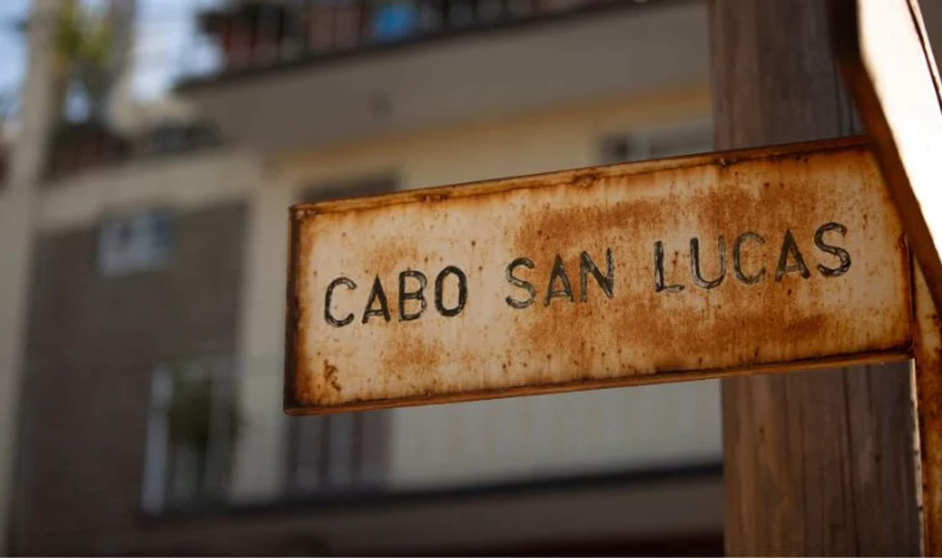 A weathered street sign reading "Cabo San Lucas" with a blurred building in the background, capturing the charm of the area.