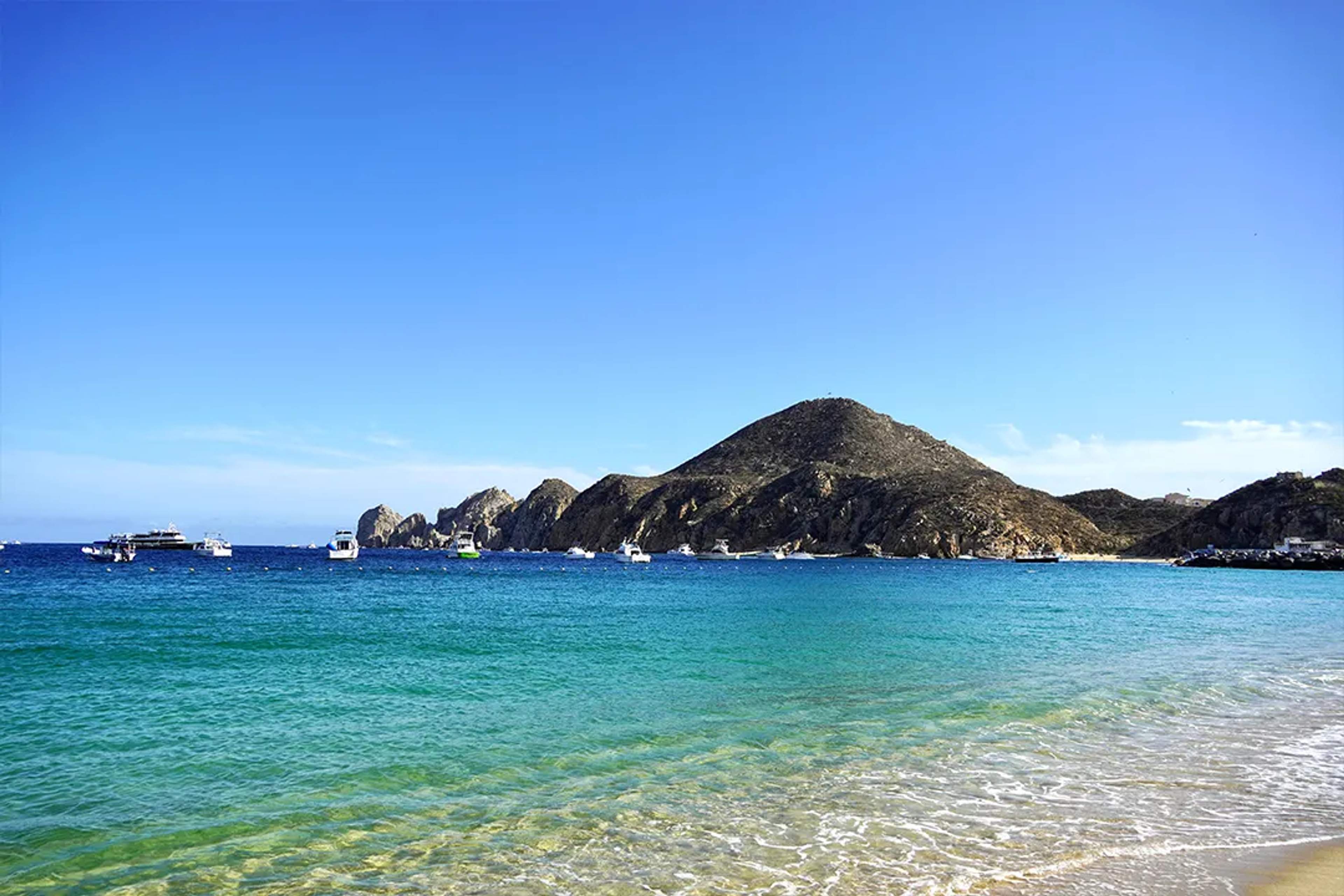 Medano Beach in Cabo San Lucas with turquoise sea, boats, and rocky hills under clear blue sky.
