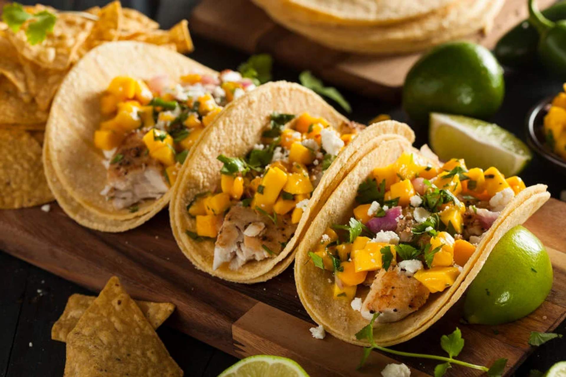 Three fish tacos topped with mango salsa and cilantro are served on a wooden board with lime wedges and tortilla chips in the background.