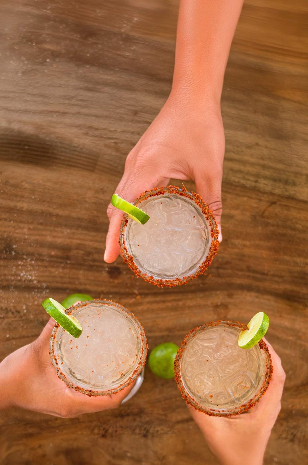 Tres manos brindando con vasos de cóctel decorados con limón y borde de chile en polvo.