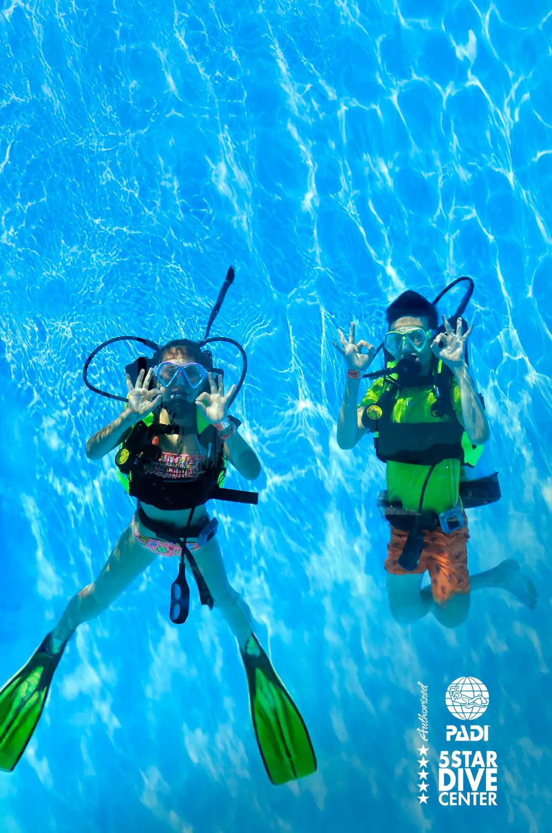 Two young divers underwater in a pool giving the "OK" hand signal, with the PADI 5 Star Dive Center logo.