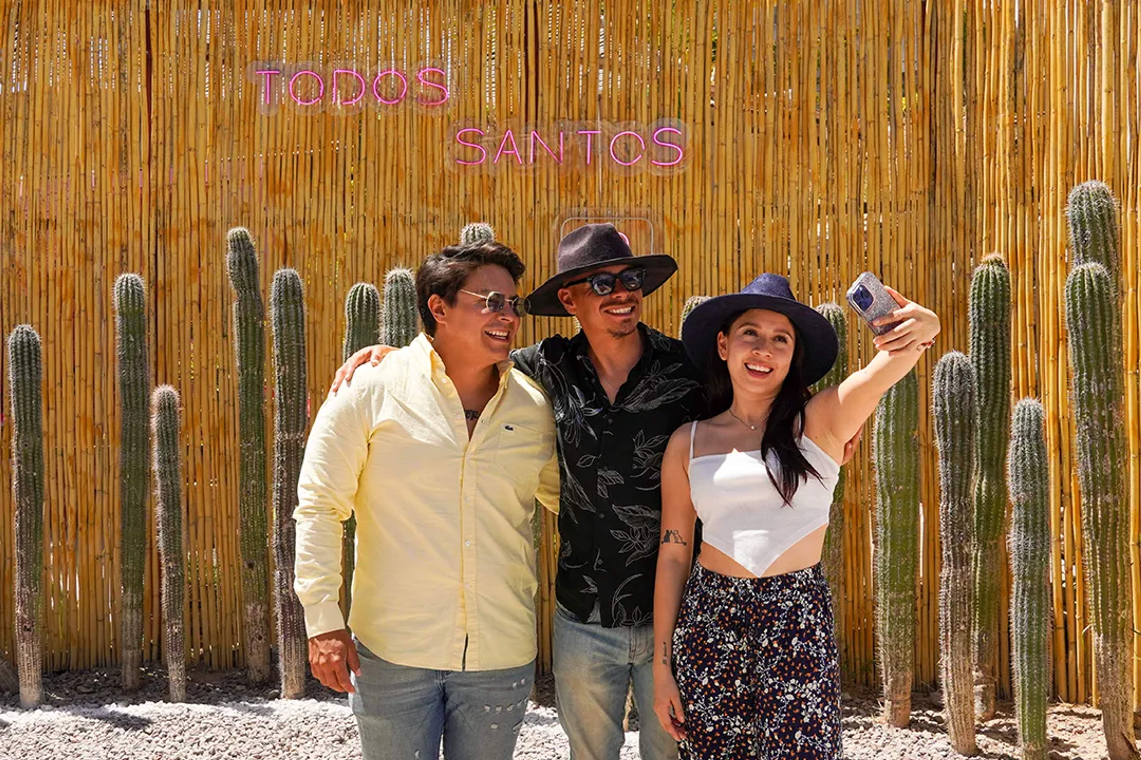 Tres amigos se toman una selfie en Todos Santos, junto a cactus y un muro de bambú al sol.