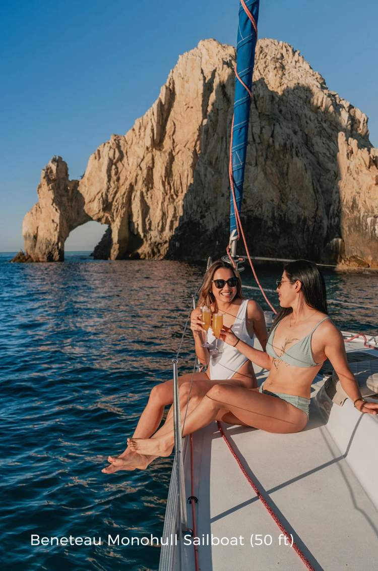 Dos mujeres brindan con bebidas en un velero cerca del arco icónico de Cabo, disfrutando de un día soleado en el mar.