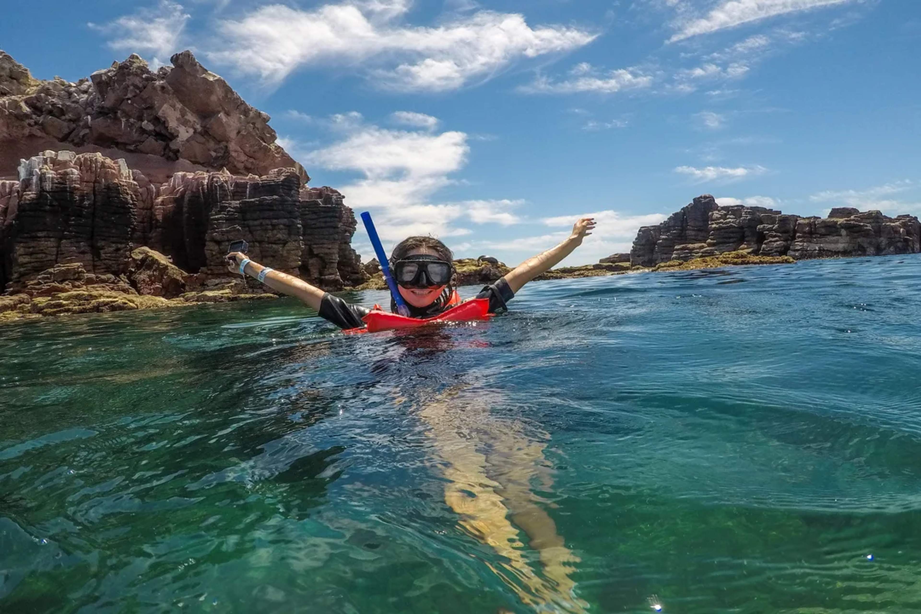 Persona practicando esnórquel en aguas claras junto a acantilados rocosos y cielo soleado.
