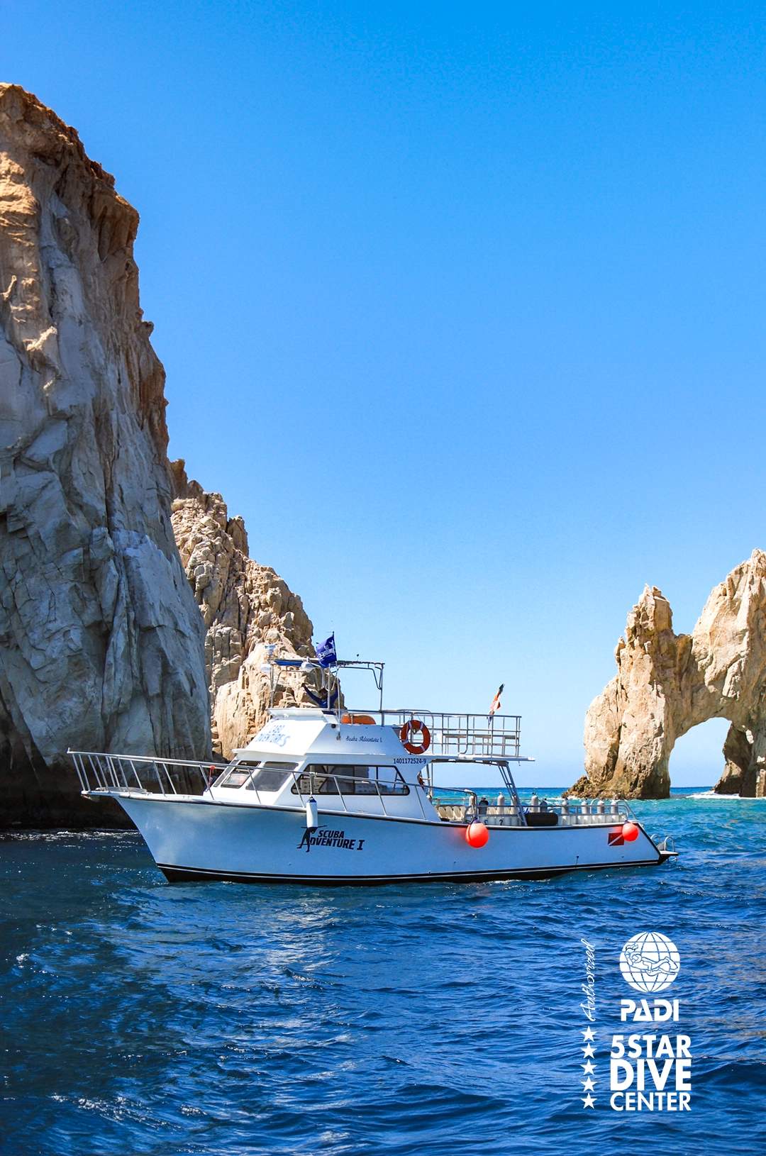 The boat "Scuba Adventure I" floats near cliffs and the iconic rock arch in Cabo San Lucas under a clear blue sky.