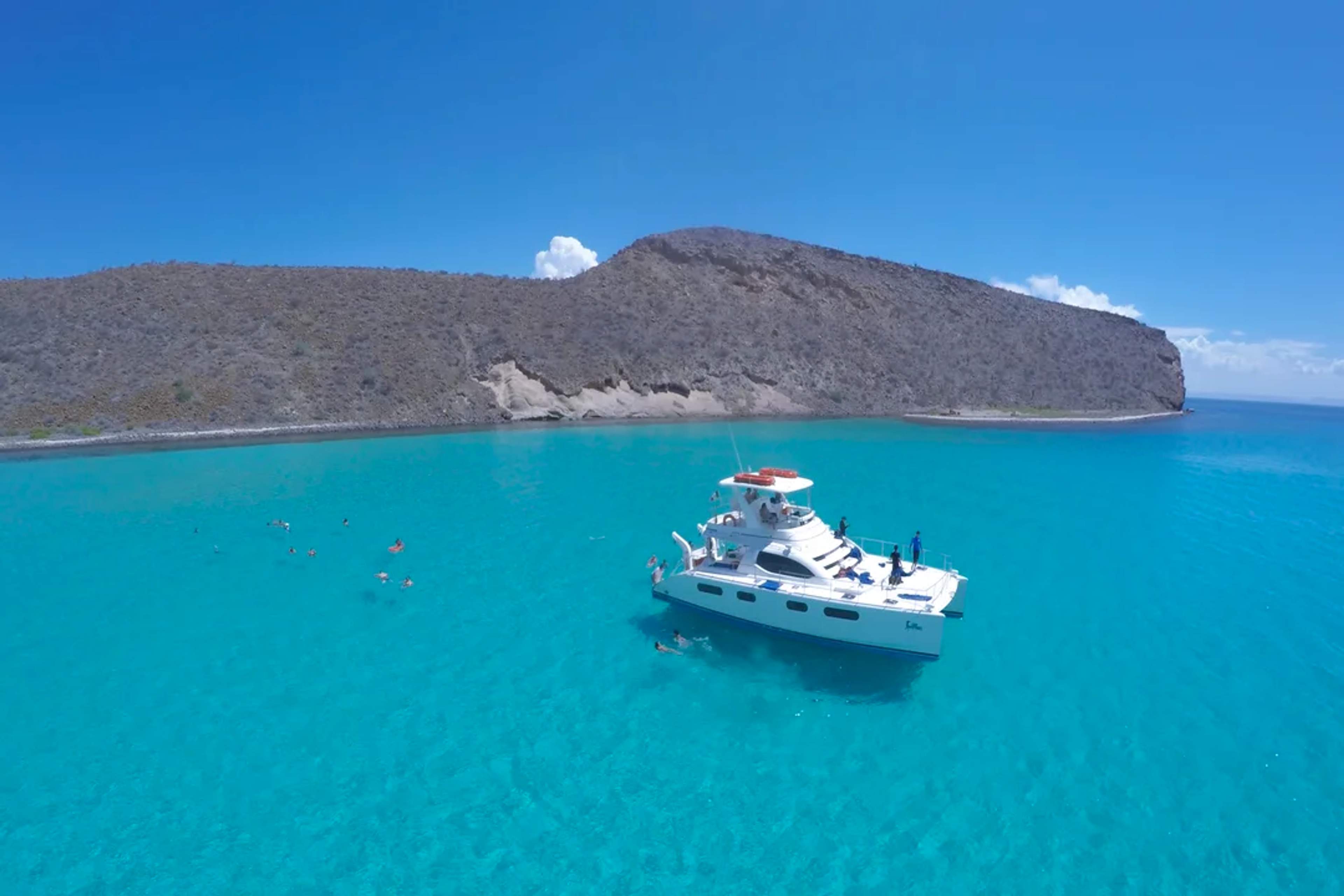 Catamarán de lujo anclado en aguas turquesa junto a un acantilado desértico, con nadadores cerca.