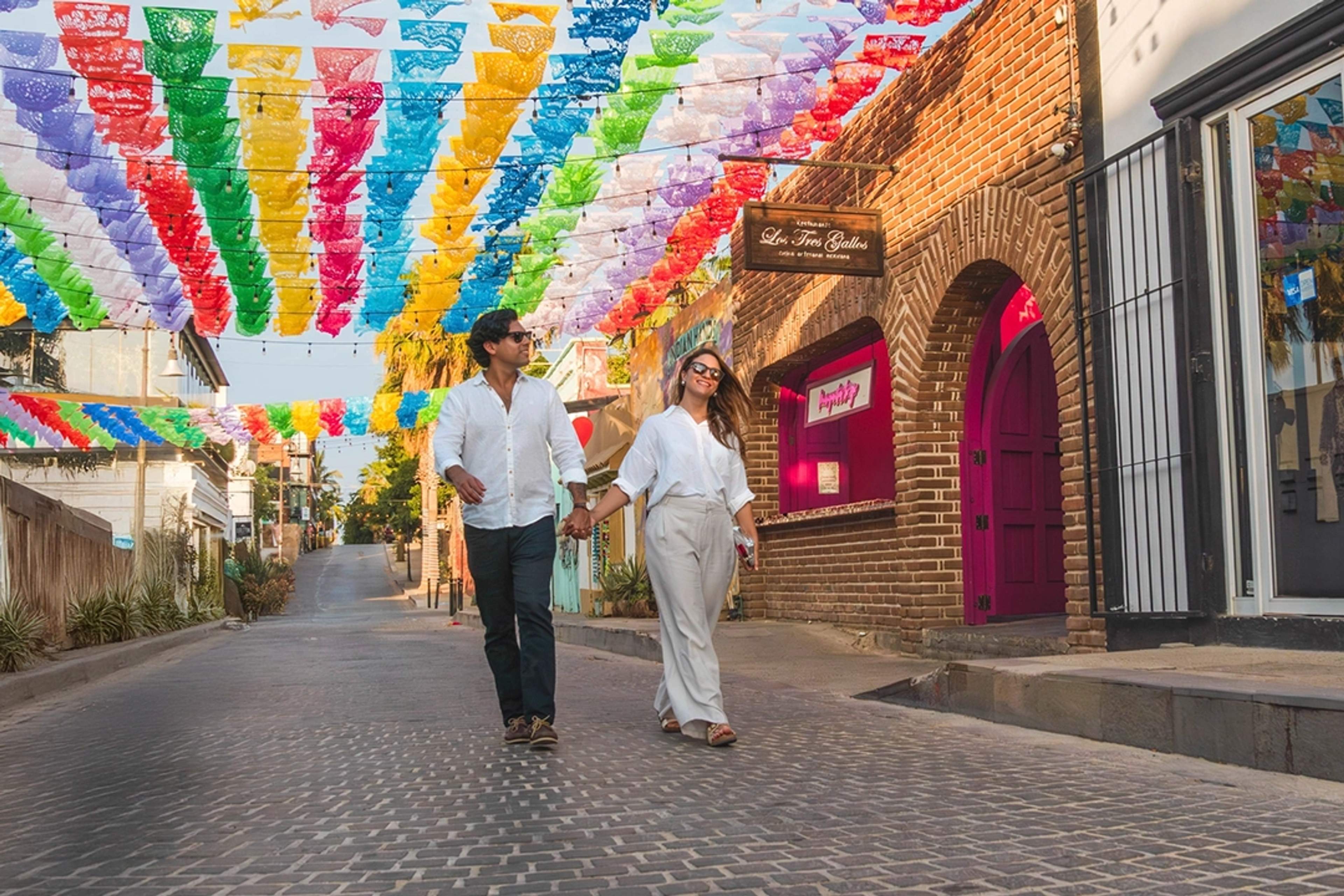 Una pareja pasea de la mano bajo coloridos banderines de papel picado en una encantadora calle empedrada de San José del Cabo.