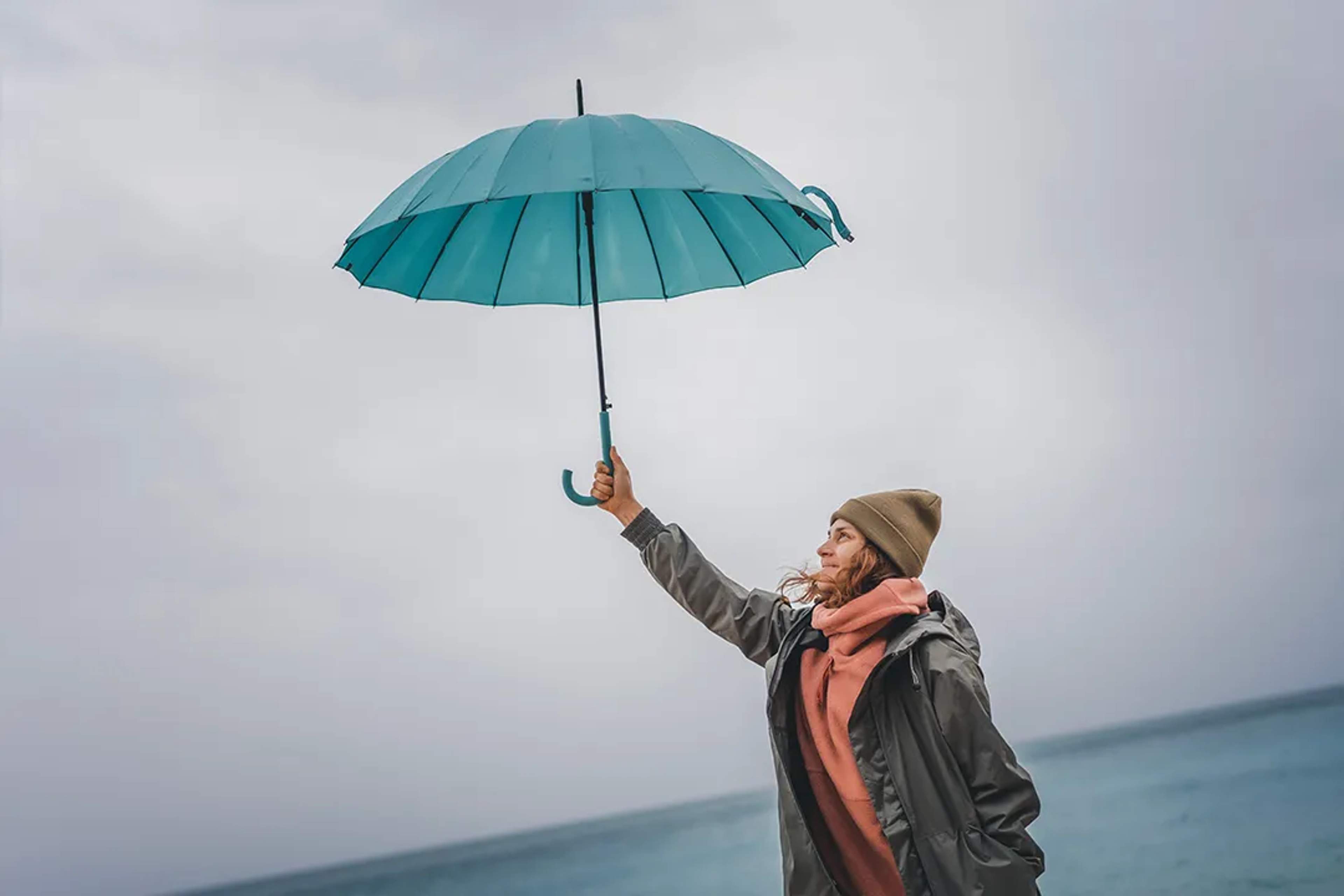 Woman holds up a teal umbrella by the sea on a windy, overcast day.