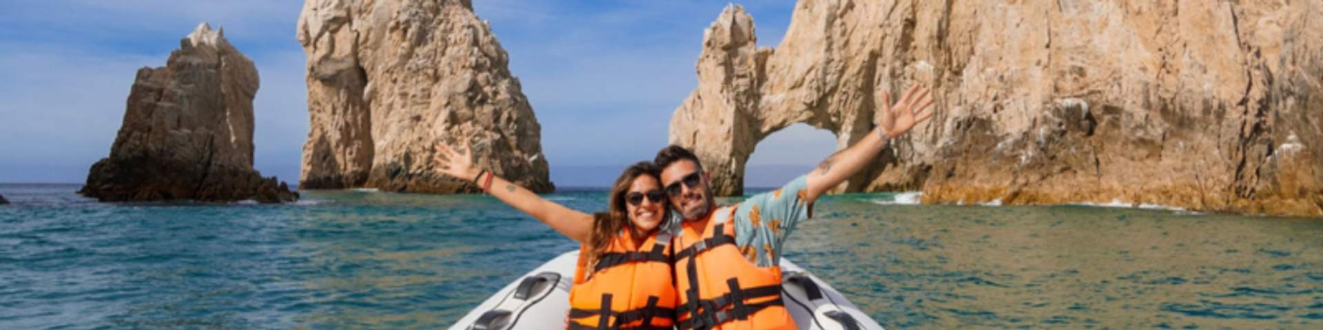 A smiling couple in orange life jackets poses with arms outstretched on a boat in front of the rock arch at Cabo San Lucas.