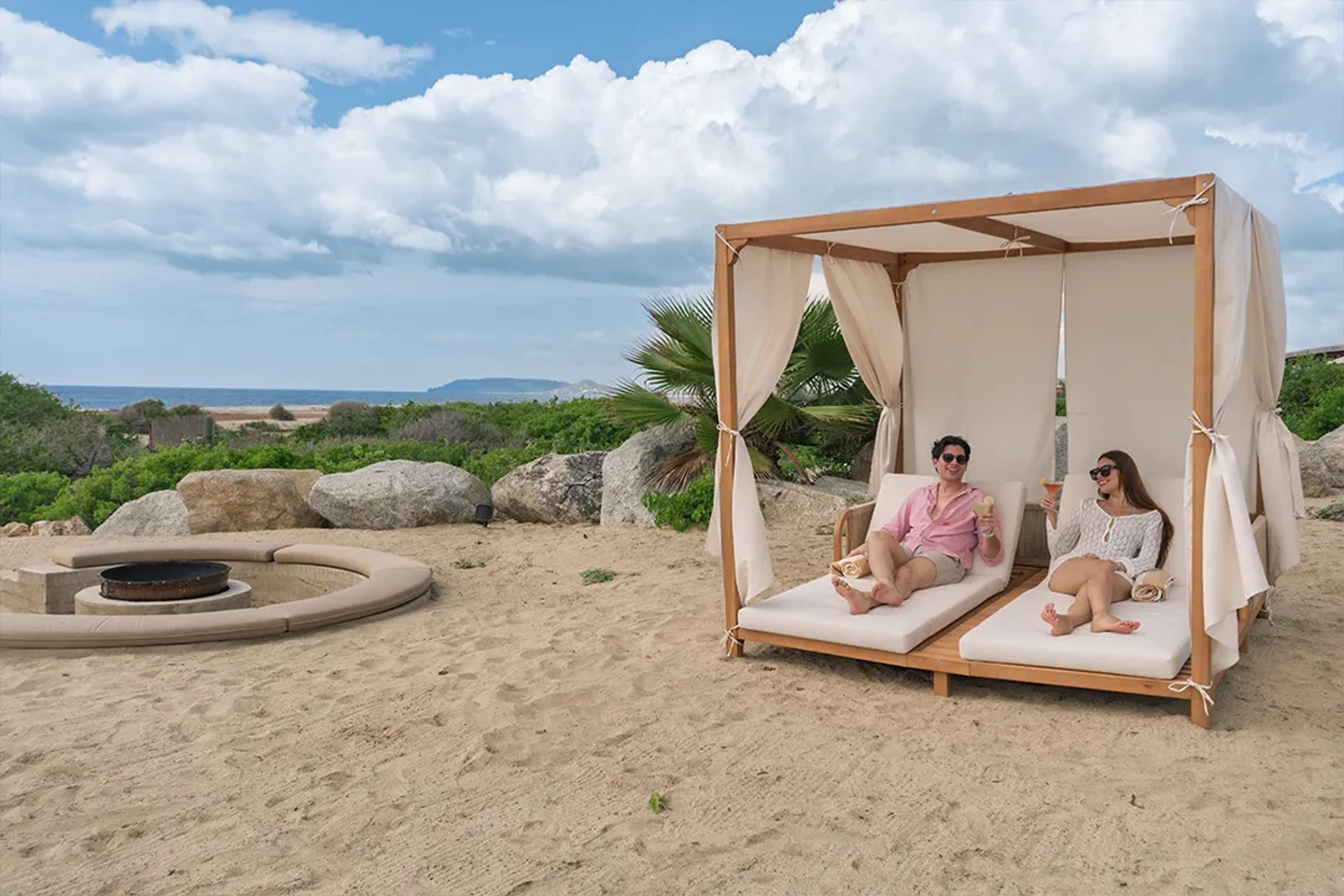 Pareja relajándose en cabaña frente al mar en Los Cabos, disfrutando bebidas y vistas al océano.