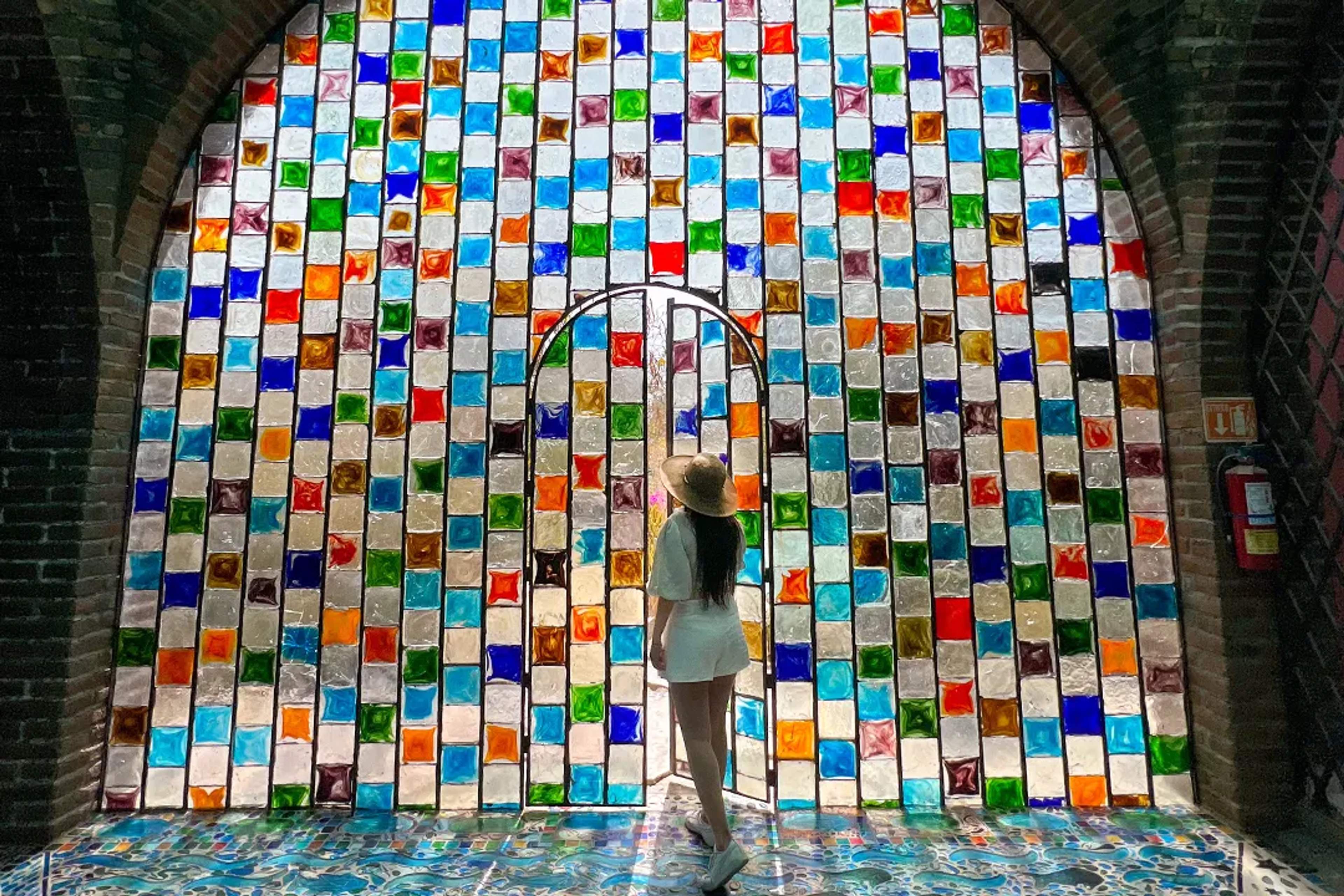 Mujer frente a un mosaico de vidrio colorido con un portón, iluminado por la luz del sol.