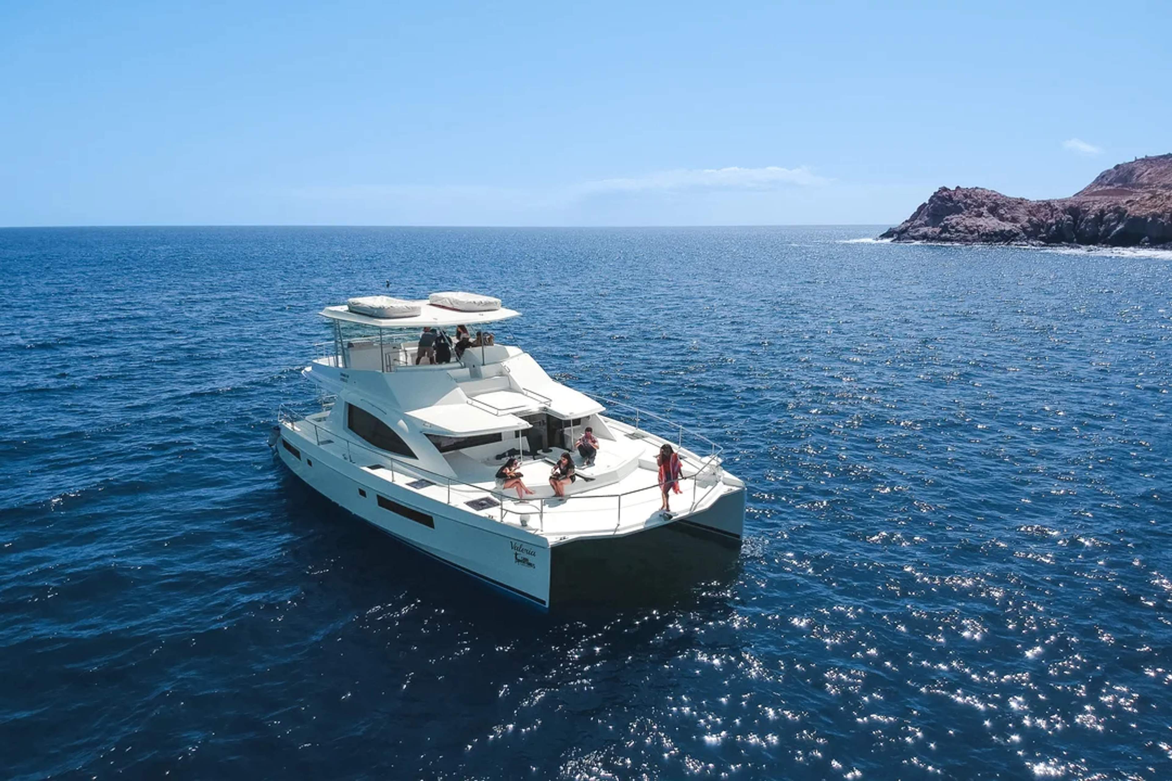 A luxury catamaran with guests enjoying the sea near a rocky coast under a bright blue sky.