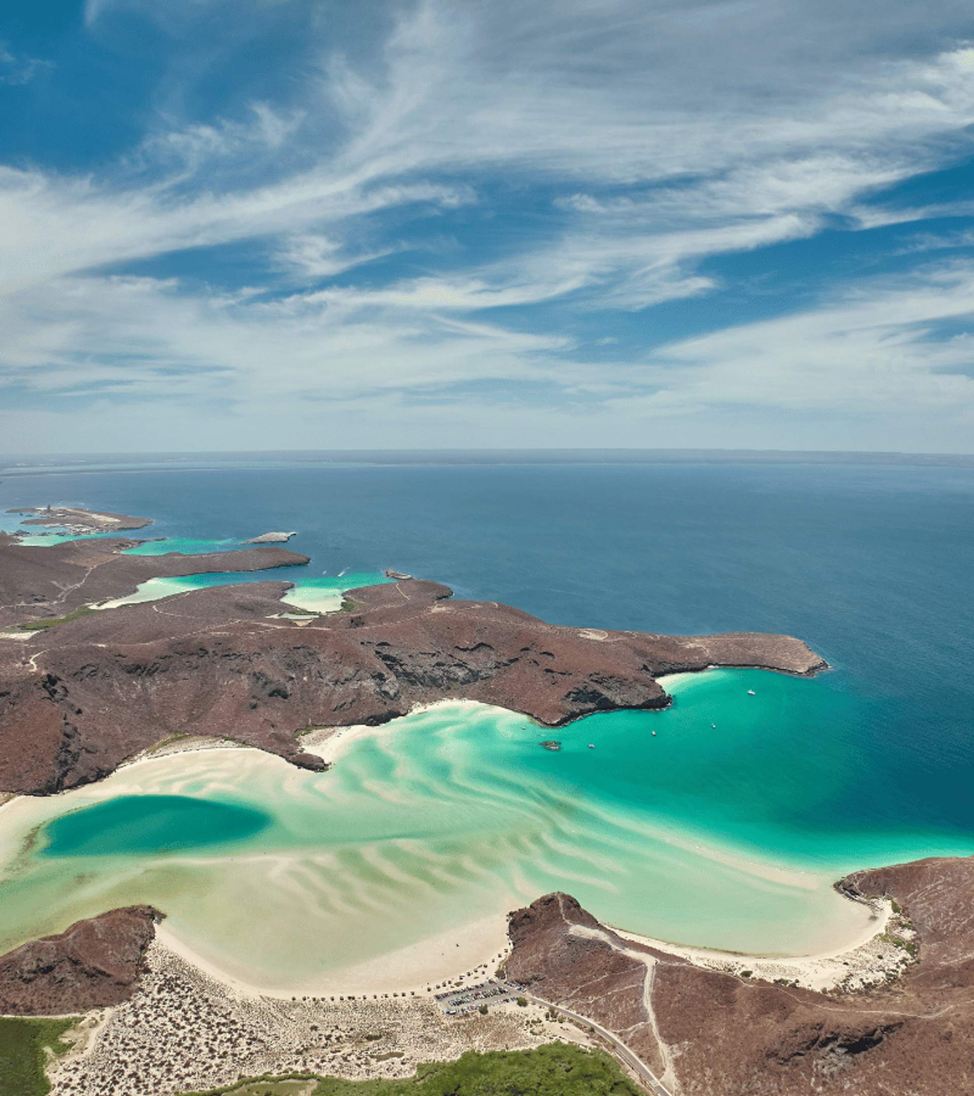 Vista aérea de una costa con varias lagunas de aguas turquesas, playas de arena blanca y colinas áridas bajo un cielo con nubes dispersas.