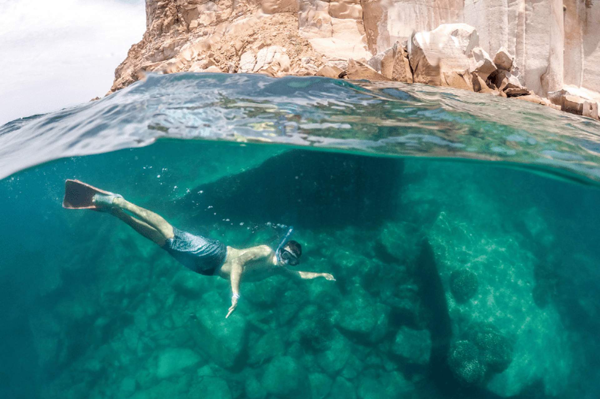 Persona haciendo snorkel en aguas cristalinas junto a una costa rocosa, con formaciones de rocas visibles bajo el agua y acantilados en la superficie.