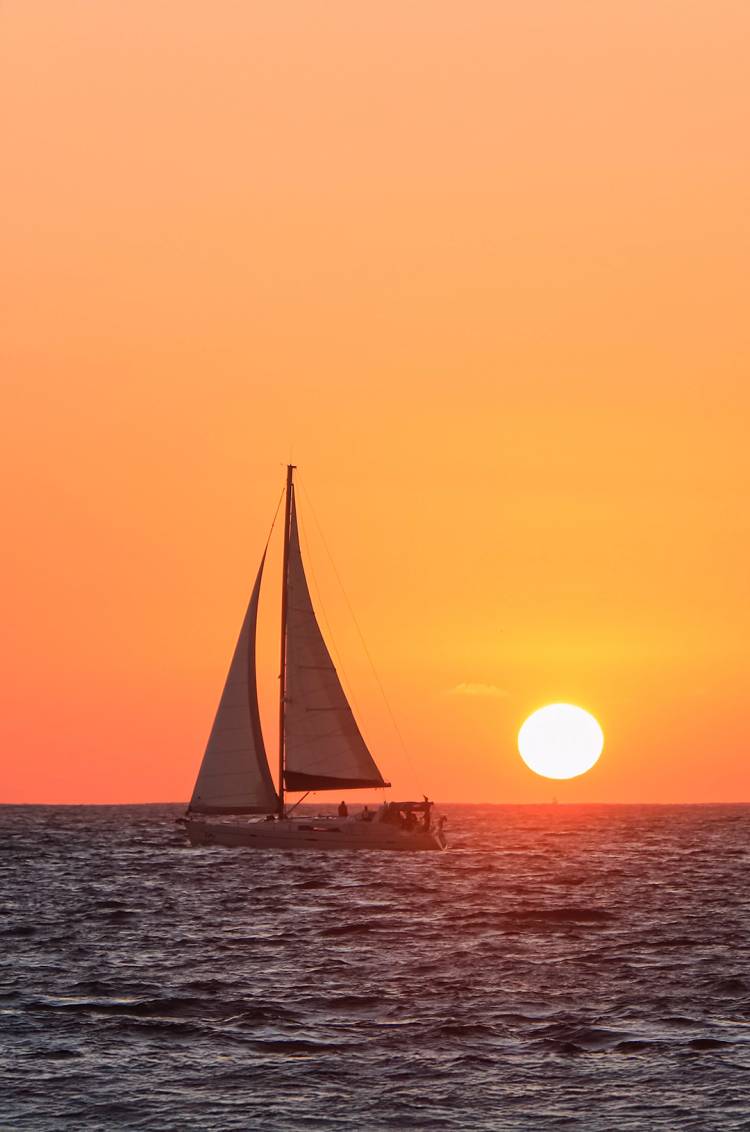 A sailboat glides across the ocean at sunset, with the glowing sun low on the horizon, creating a serene and golden scene.