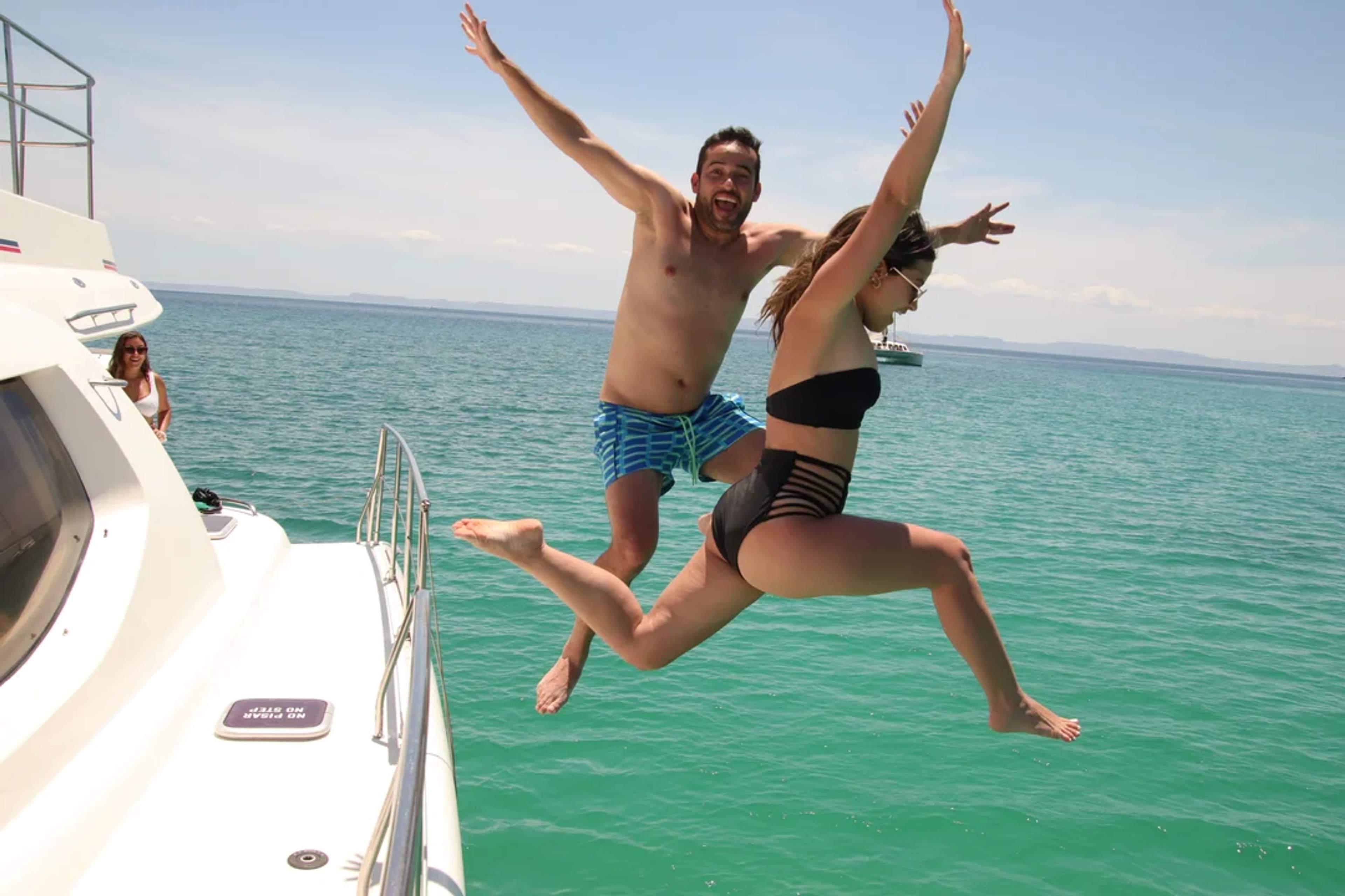 Una pareja salta con alegría desde un bote al mar turquesa, disfrutando un día soleado perfecto.