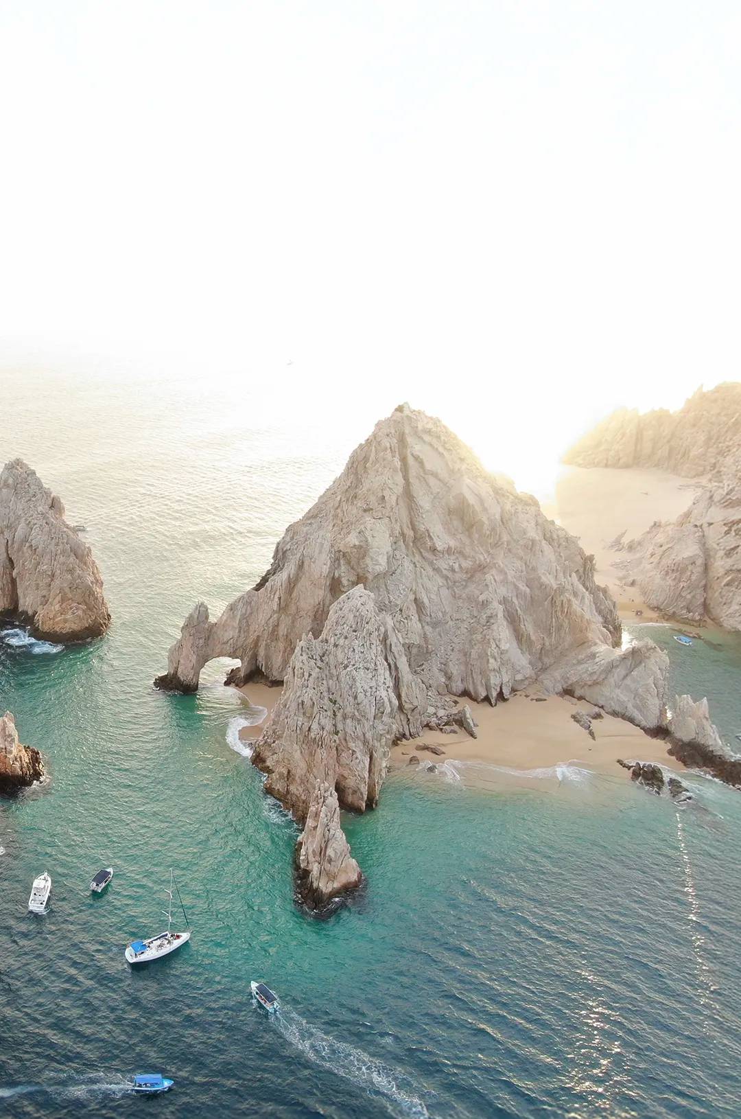 Aerial view of El Arco in Los Cabos with boats on turquoise water at sunset.