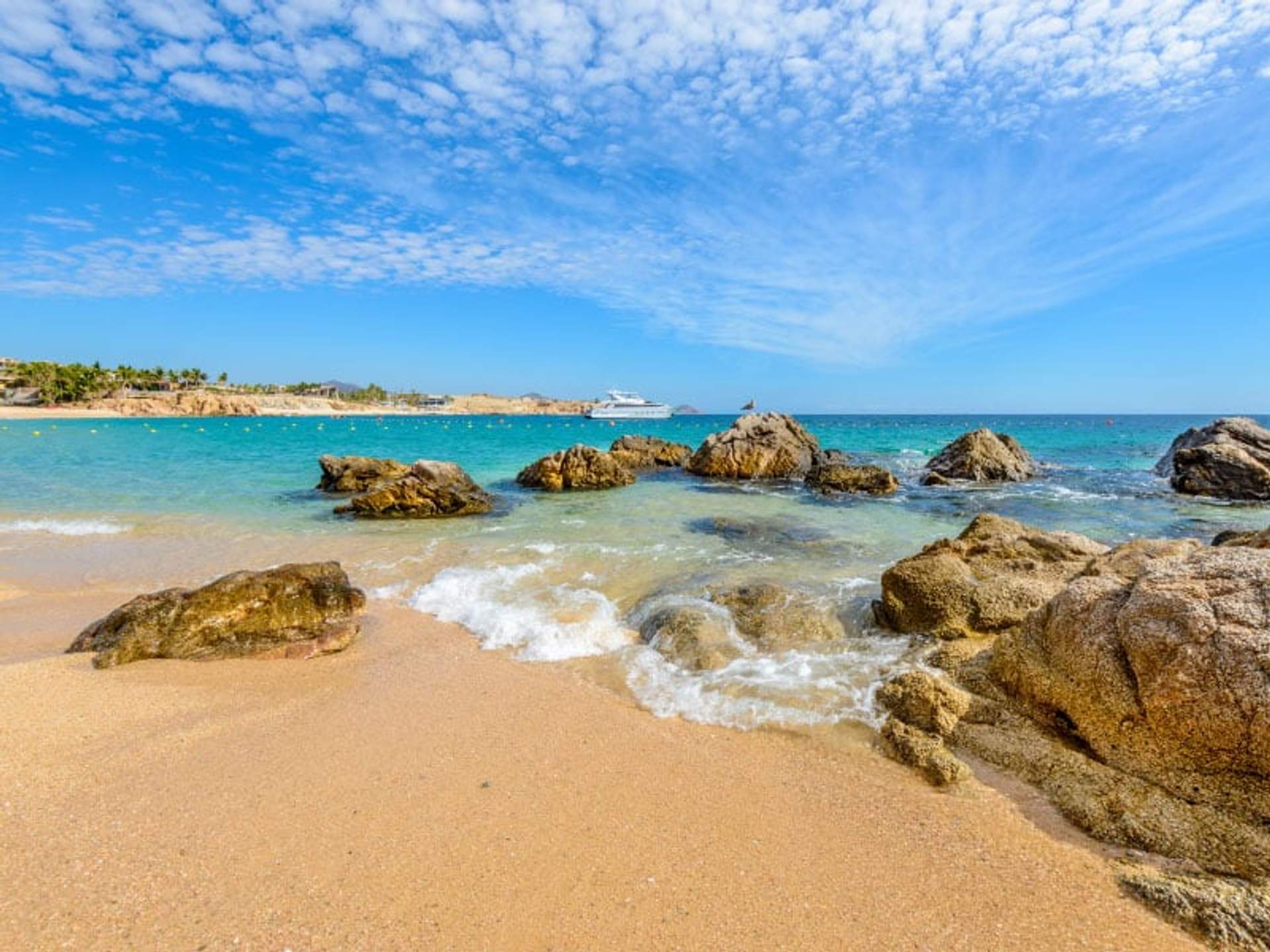Playa con rocas y arena dorada, aguas turquesas y un barco en la distancia bajo un cielo azul.