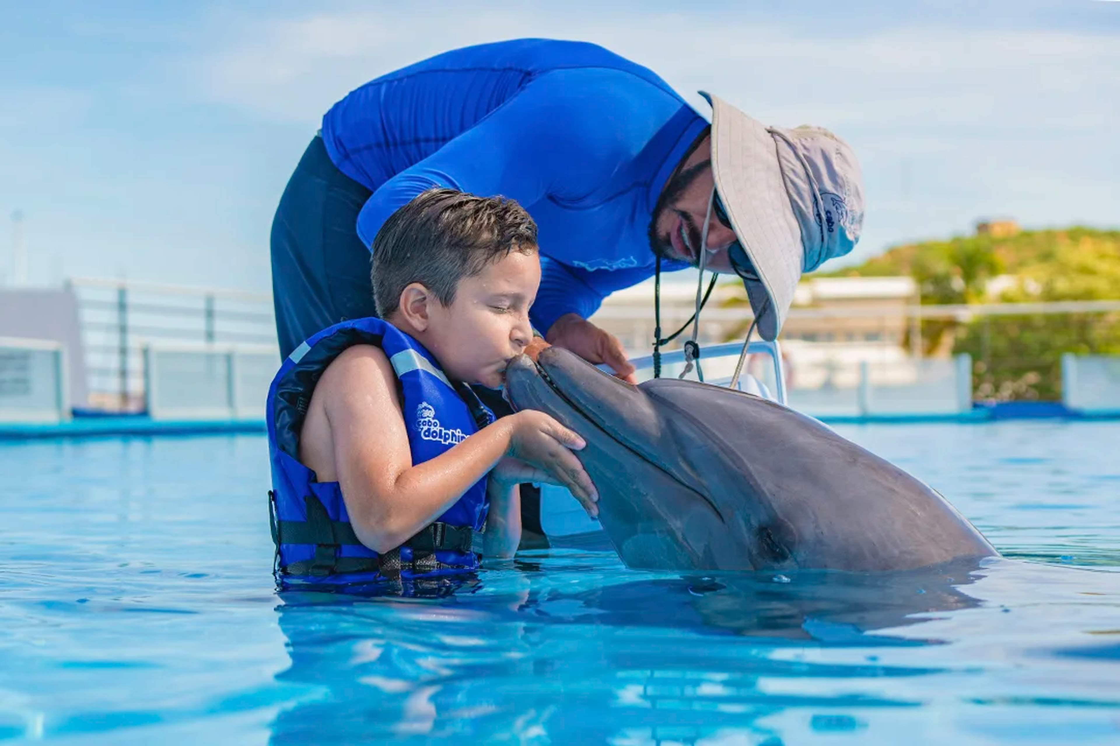 A young boy wearing a life jacket kisses a dolphin in a pool, guided by an instructor in a blue shirt and sun hat.