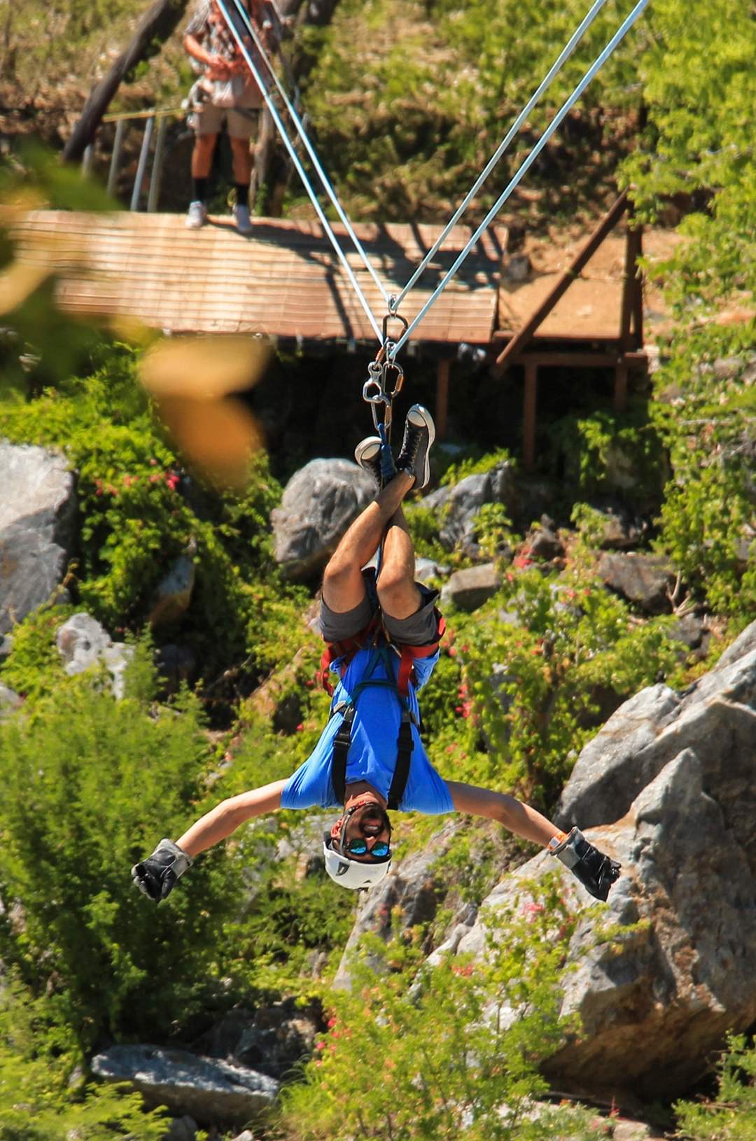 A person zip-lining upside down over a rocky forest area, wearing a blue shirt, helmet, and harness.