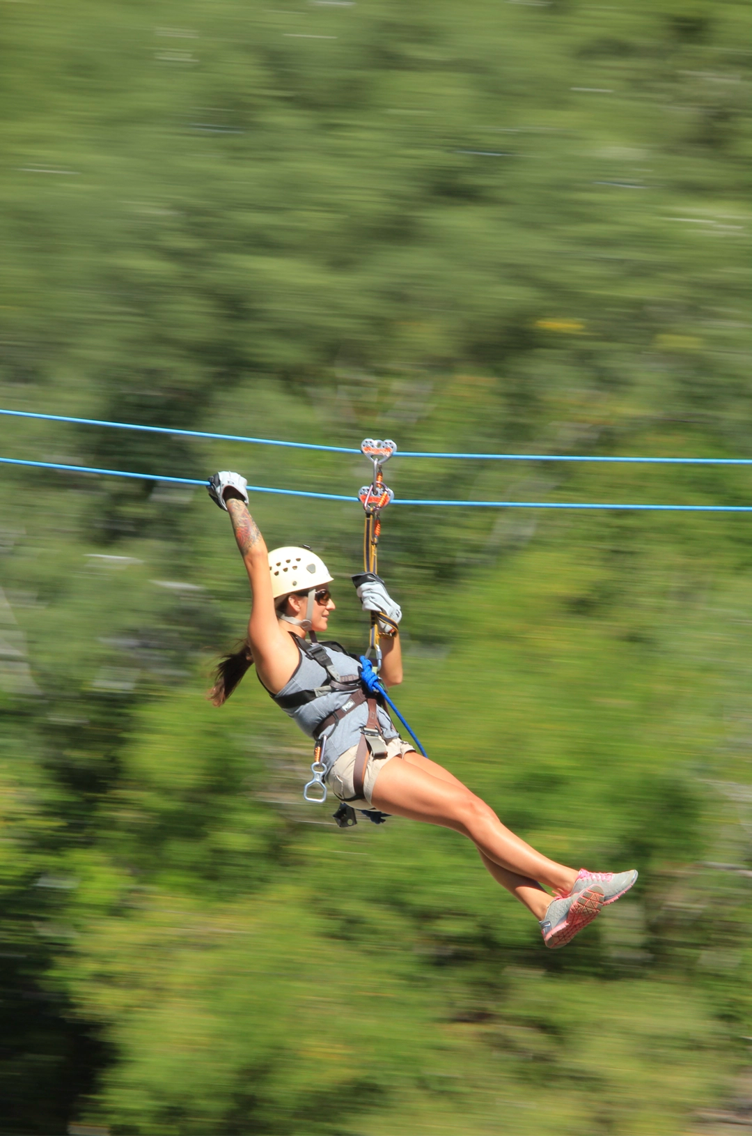 A woman zip-lining through a forest, wearing a helmet, harness, and gloves, smiling as she speeds along.
