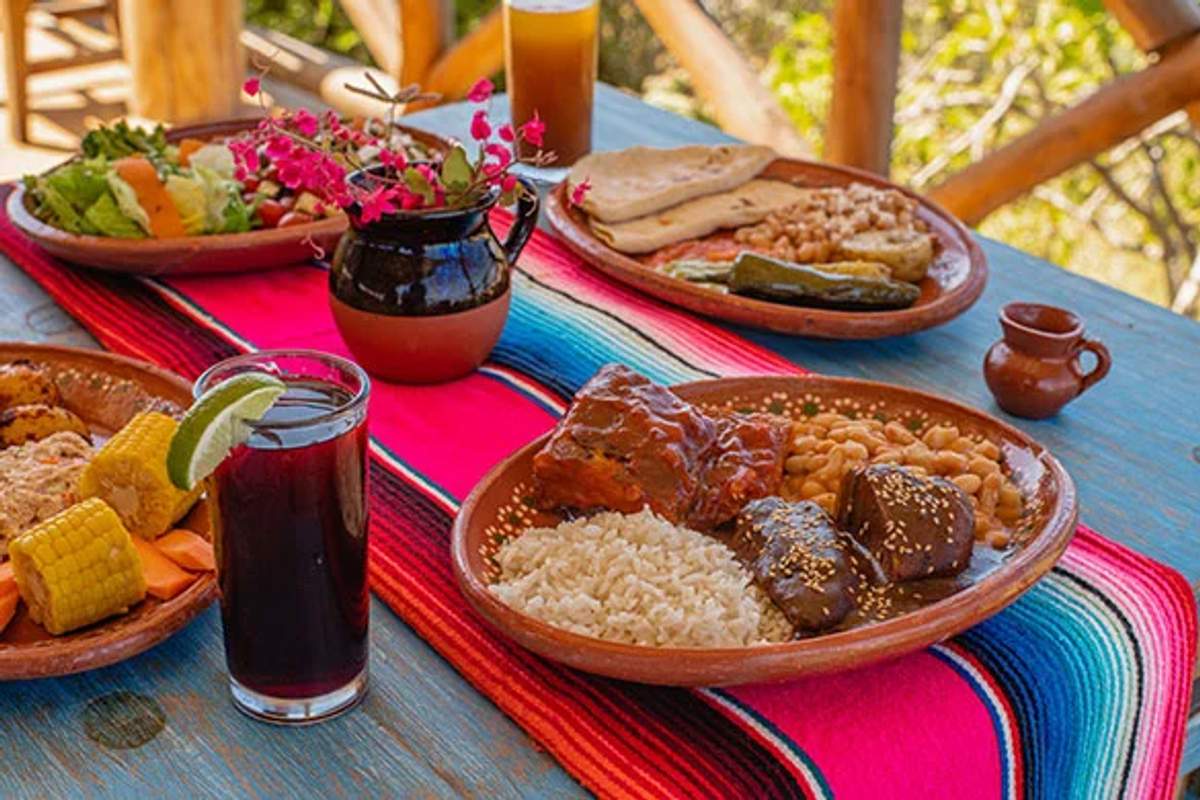 A traditional Mexican meal on a colorful tablecloth, featuring rice, beans, meat, corn, tortillas, and drinks, set on a rustic wooden table.