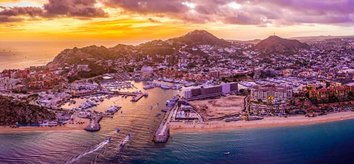 An aerial view of Cabo San Lucas at sunset, showcasing the marina filled with boats and the vibrant cityscape nestled between hills. The golden and purple hues of the sky reflect on the calm ocean waters, enhancing the picturesque beauty of this popular destination.