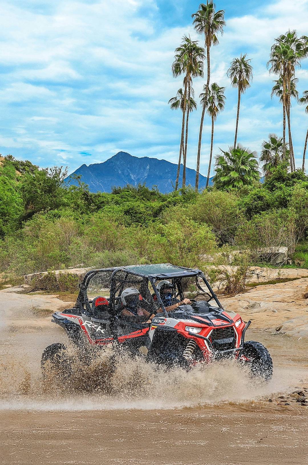 Two people drive a UTV through a muddy puddle, splashing water, with tall palm trees and a mountain backdrop.