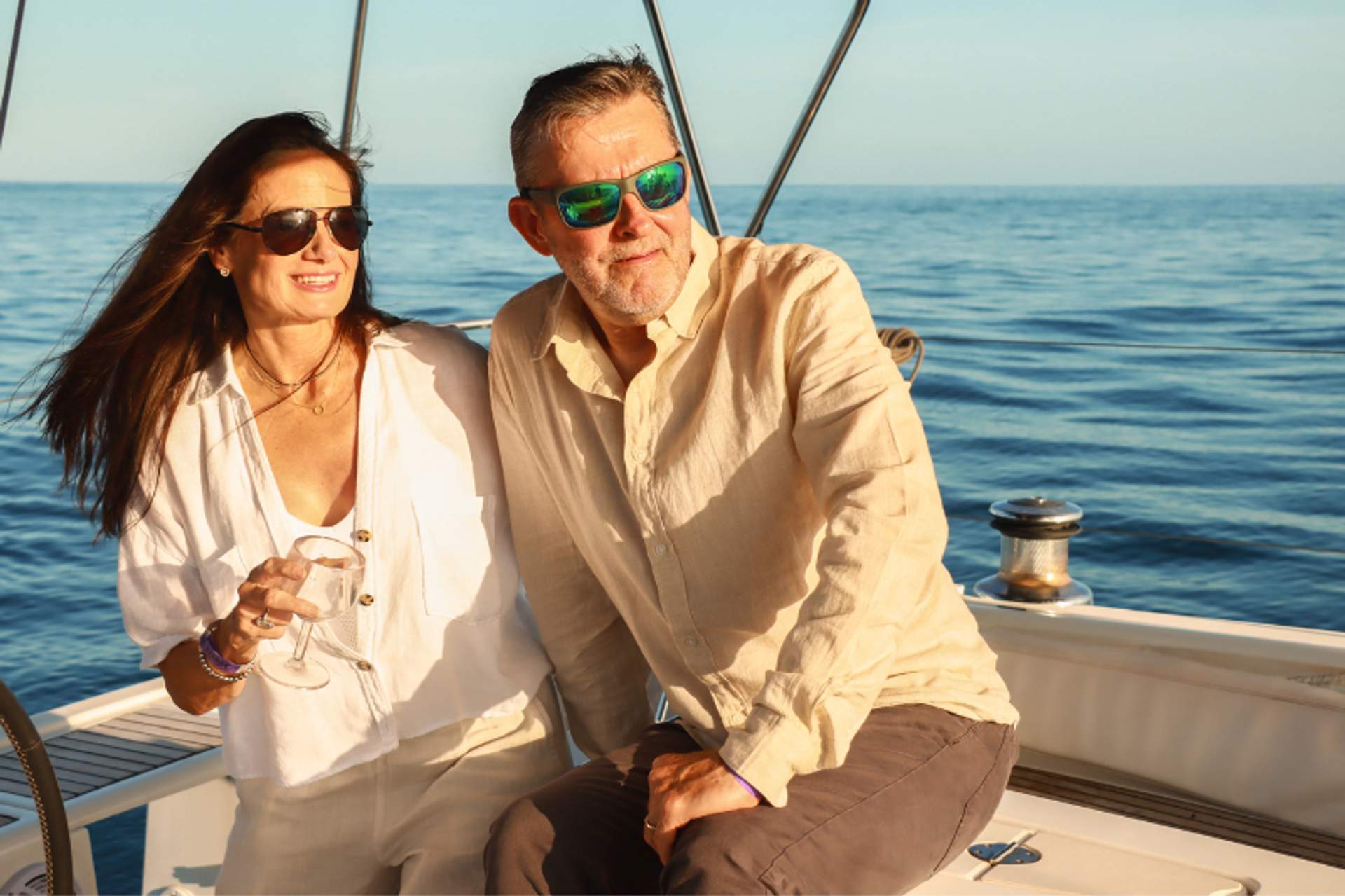A couple enjoys a sunny day on a boat, both wearing sunglasses and casual, light-colored clothing. The man sits on the boat's edge, while the woman stands beside him holding a glass, with the open sea and clear sky in the background, creating a serene and relaxed atmosphere.