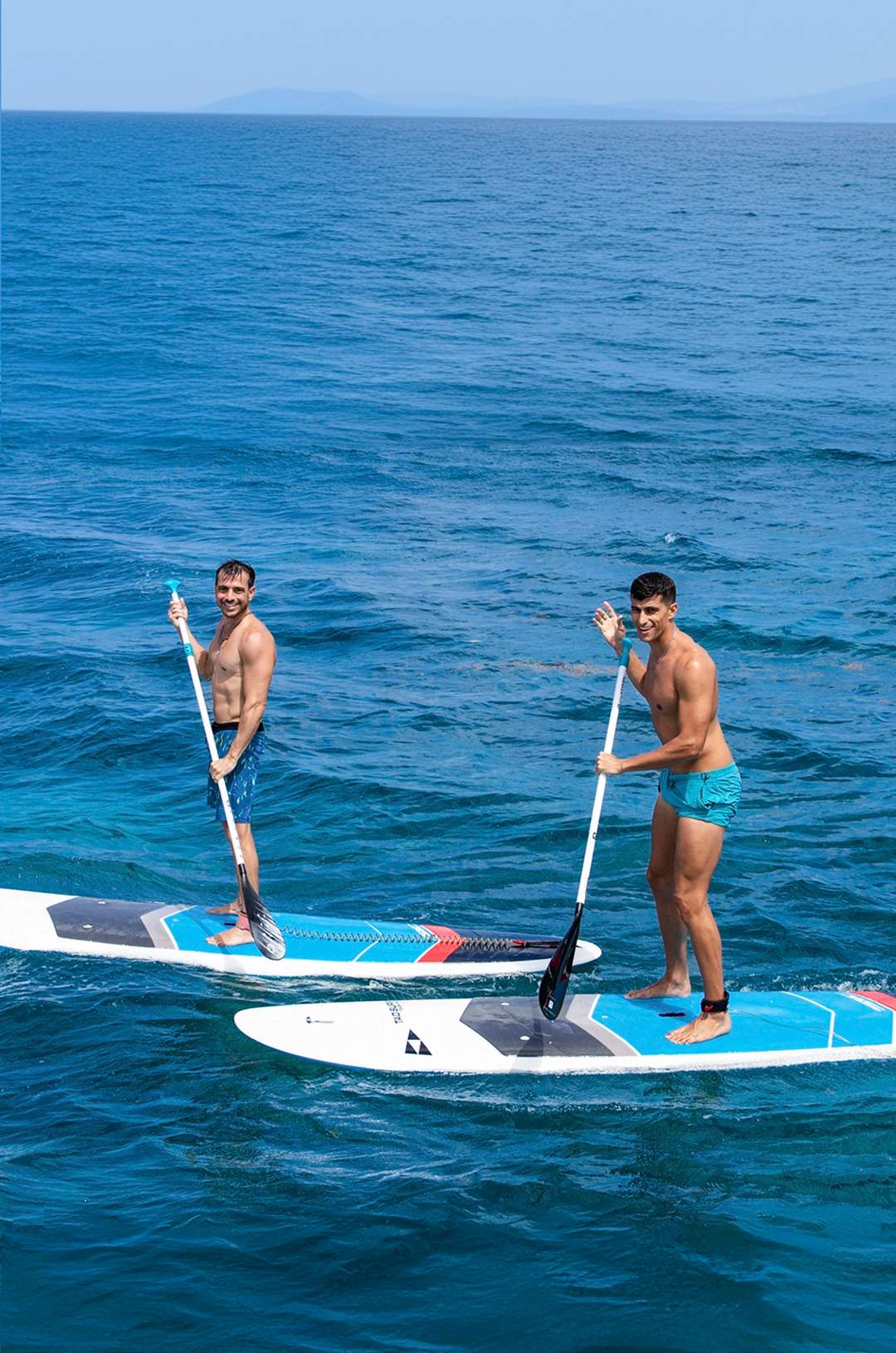 Two men paddleboarding on clear blue ocean water, smiling and waving at the camera.