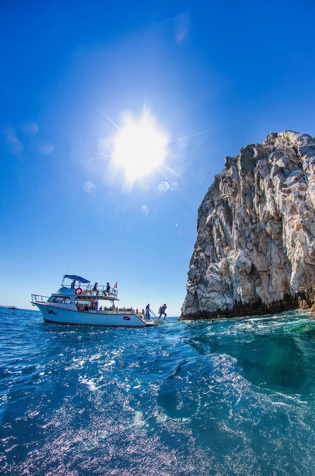 A boat anchored near a rocky cliff with a diver jumping into the clear blue ocean under a bright sunlit sky.