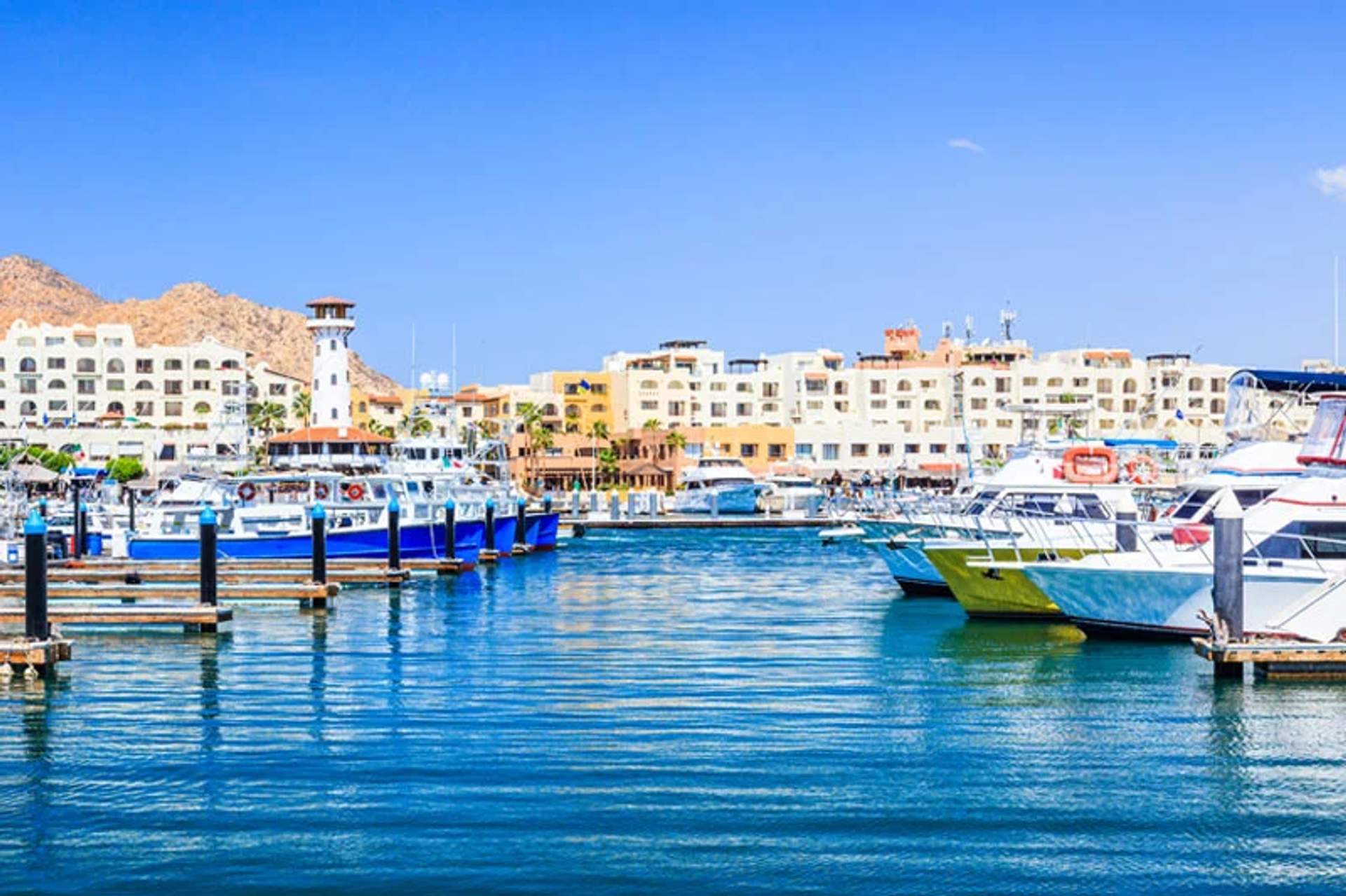 Harbor with numerous boats docked on a sunny day, with multi-story buildings and hills in the background. The water reflects the bright colors of the boats and the clear blue sky.