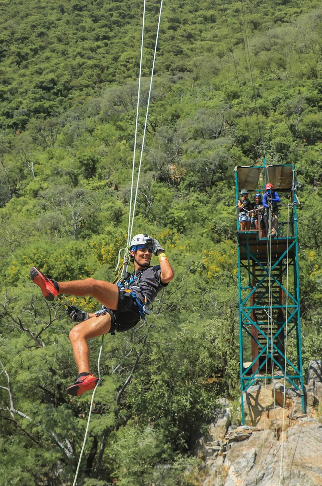 A person zip-lining through a forested area with a tall platform in the background, wearing a helmet and harness.