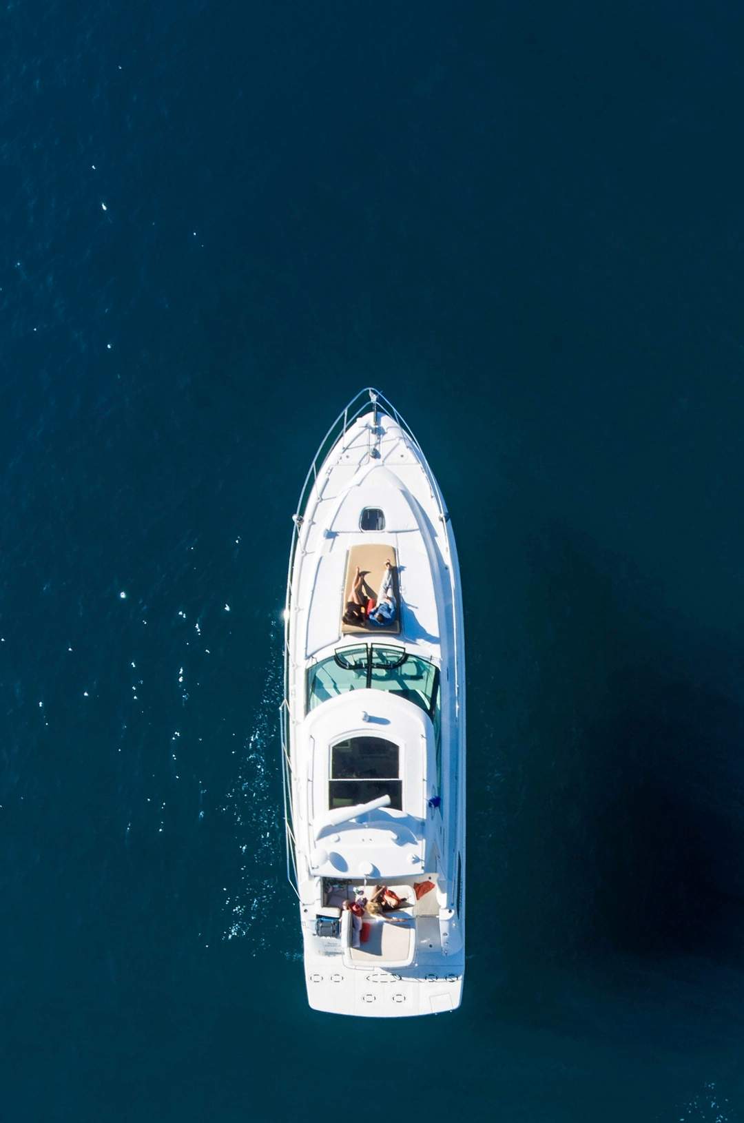Aerial view of a white yacht cruising on deep blue water, with people relaxing on the deck, enjoying the sun and sea.