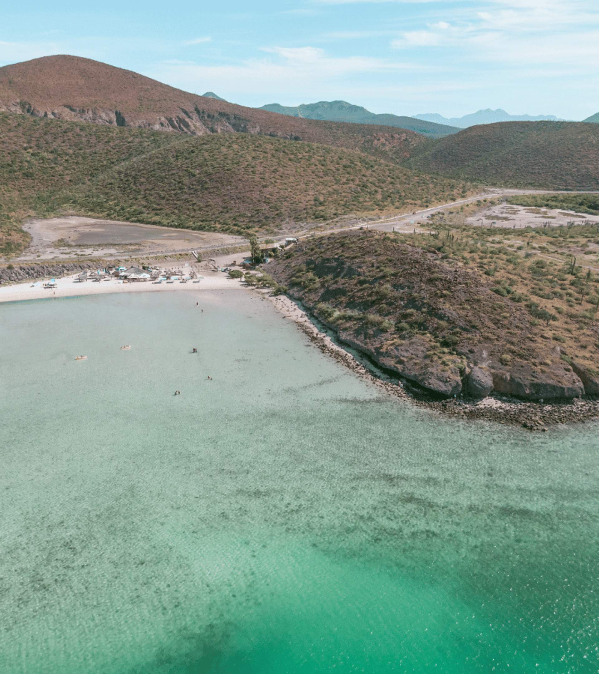 Aerial view of a serene beach with clear turquoise waters, small groups of people, rocky hills and an arid landscape under a bright sky.