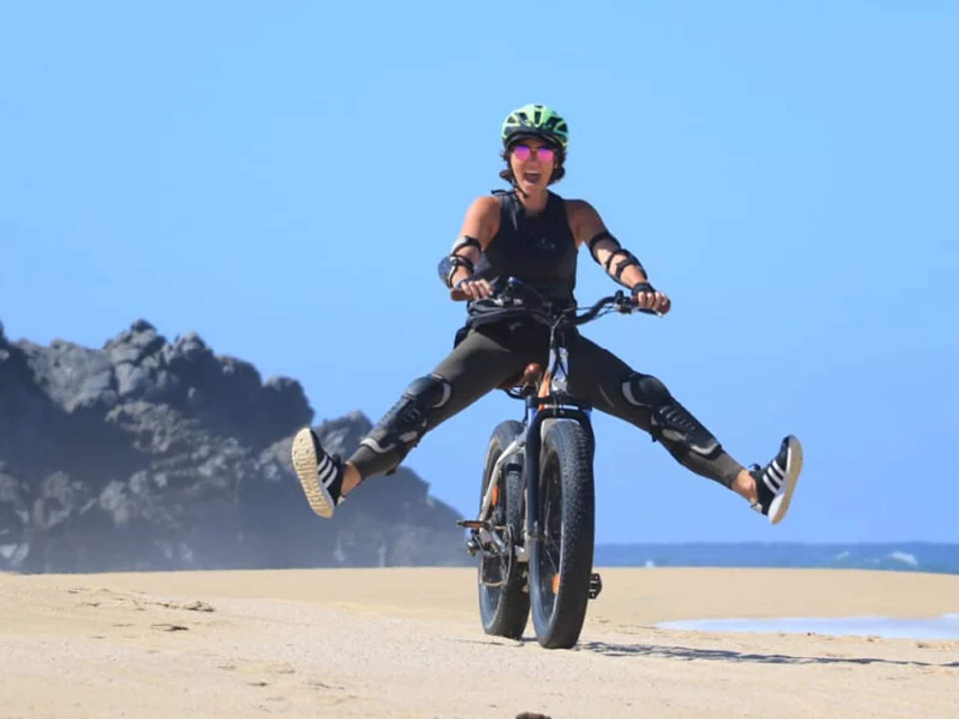 A person wearing protective gear rides an electric bike on the beach, lifting both legs in the air while smiling.