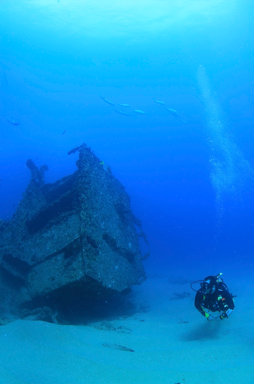 Three scuba divers explore a shipwreck on the ocean floor, surrounded by blue water and scattered debris.
