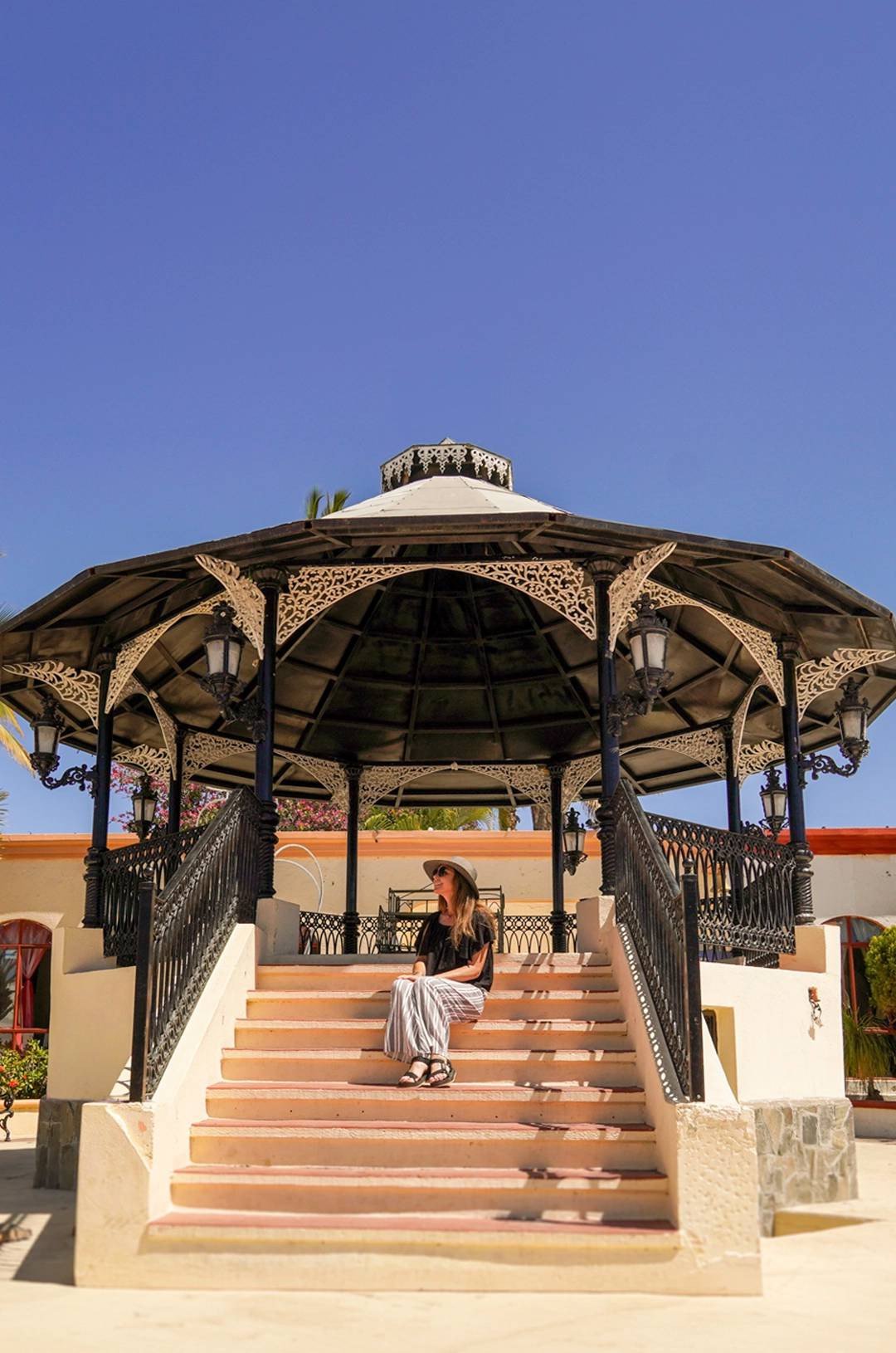 Woman in a hat sitting on the steps of an ornate gazebo under a clear blue sky.