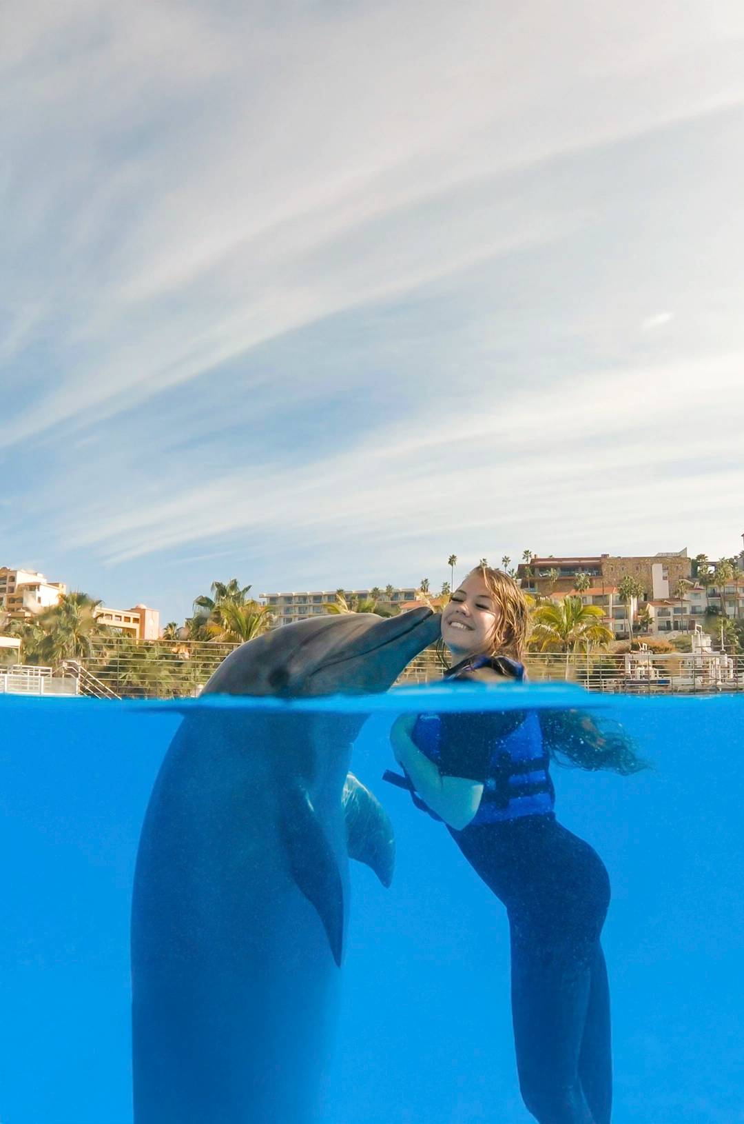 Smiling woman in a life jacket receiving a kiss from a dolphin in clear water, with a resort in the background.