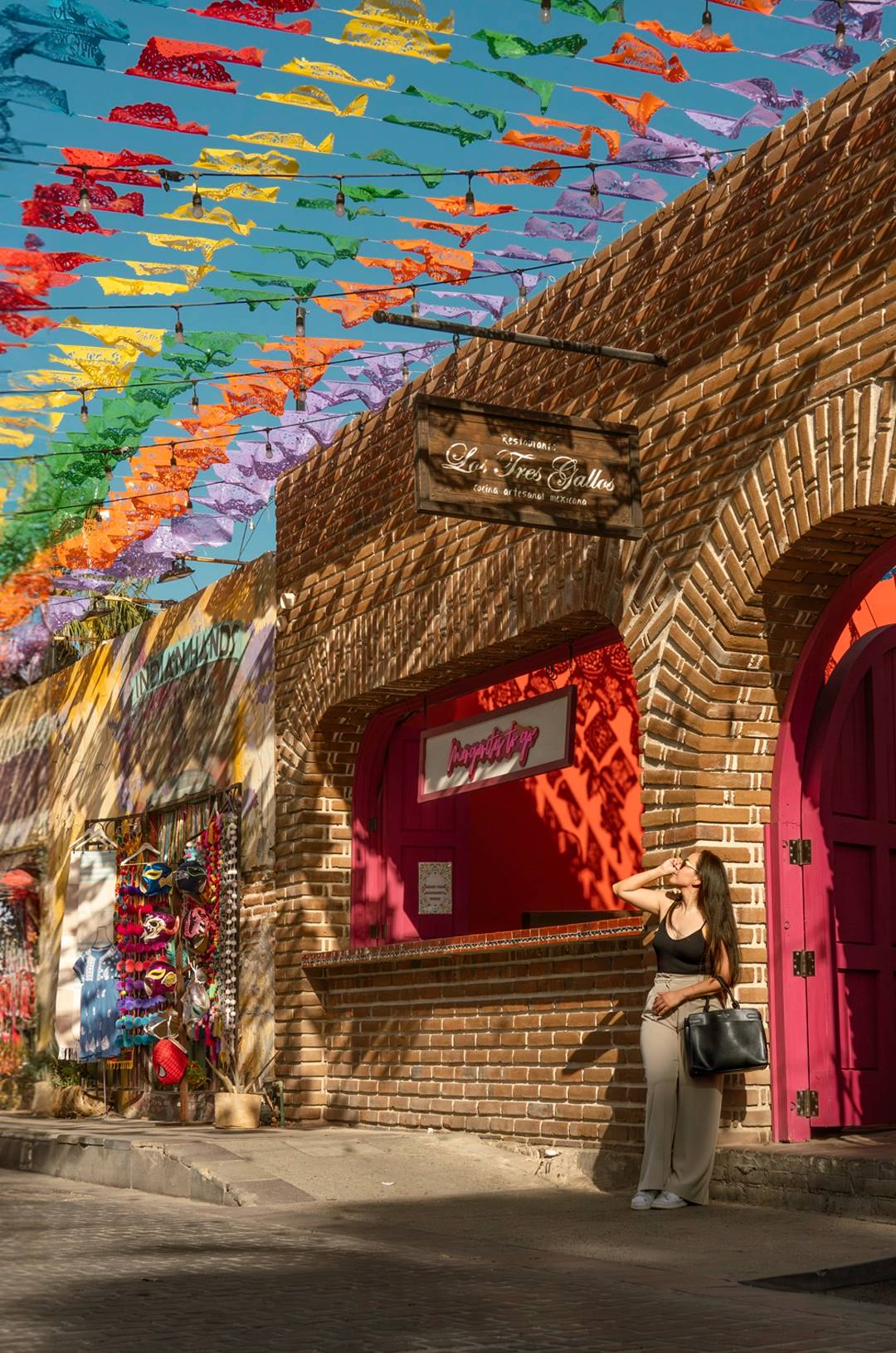 A woman stands in front of a colorful shop in San José del Cabo, under a sky decorated with vibrant papel picado banners.