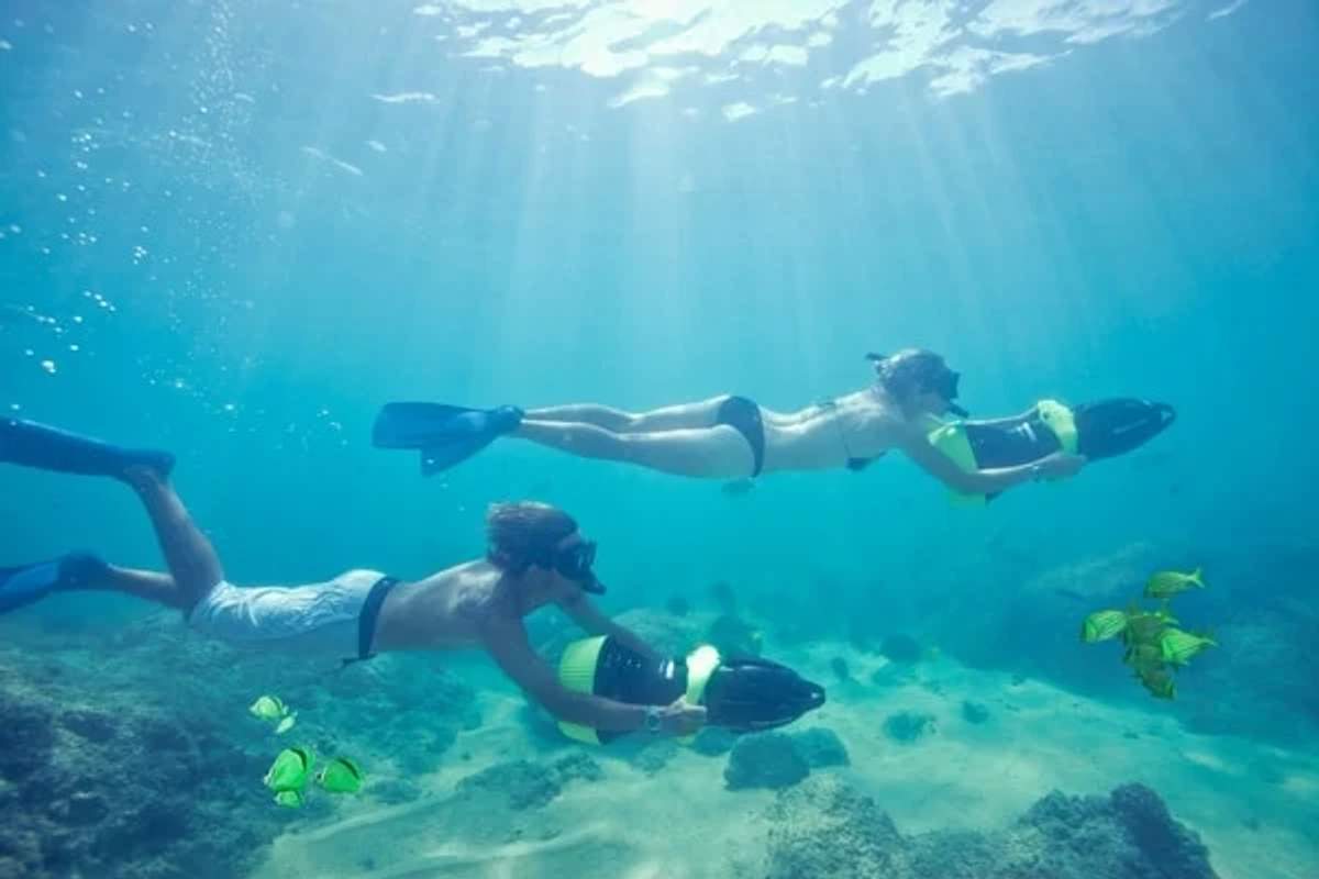 Two snorkelers, one male and one female, glide underwater with sea scooters in a clear blue sea, surrounded by small yellow fish and a rocky seabed. Sunlight filters down through the water, creating a serene and vibrant underwater scene.