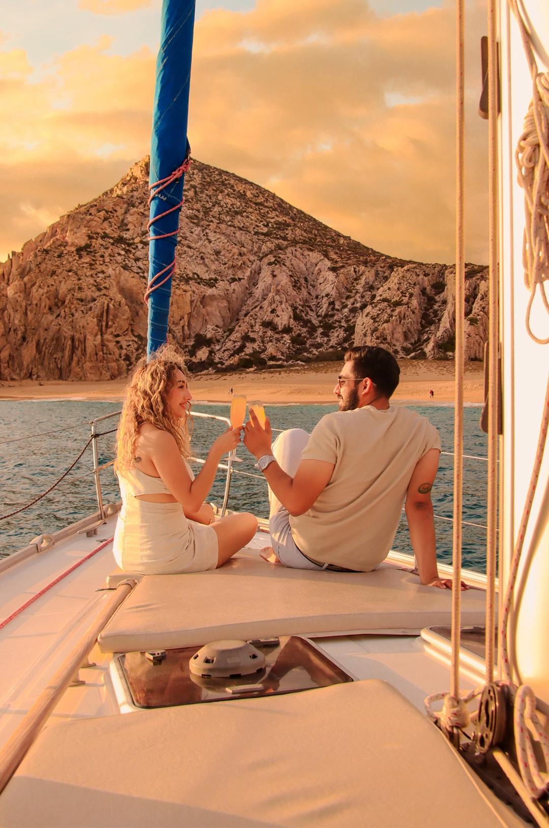 Couple toasting with drinks on a sailboat in Cabo, enjoying a scenic view of rocky cliffs at sunset.