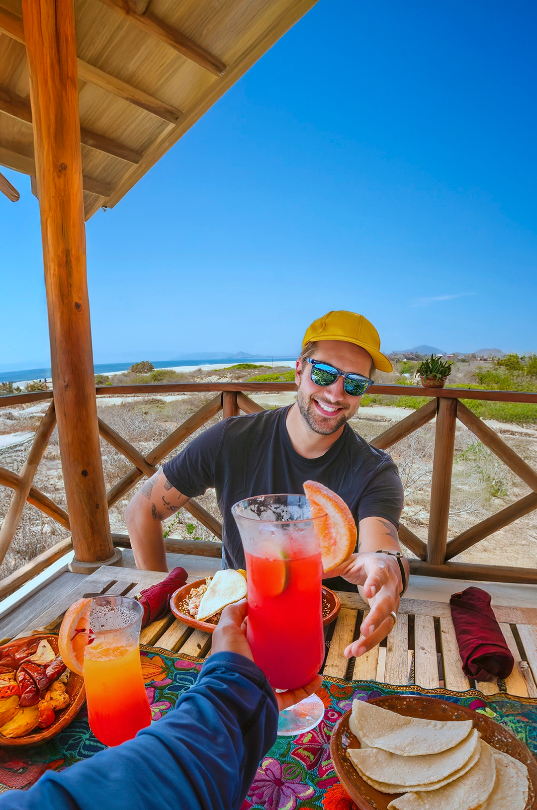 Two people sit on colorful blankets, watching the sunset over the ocean with snacks and drinks nearby on a sandy beach.