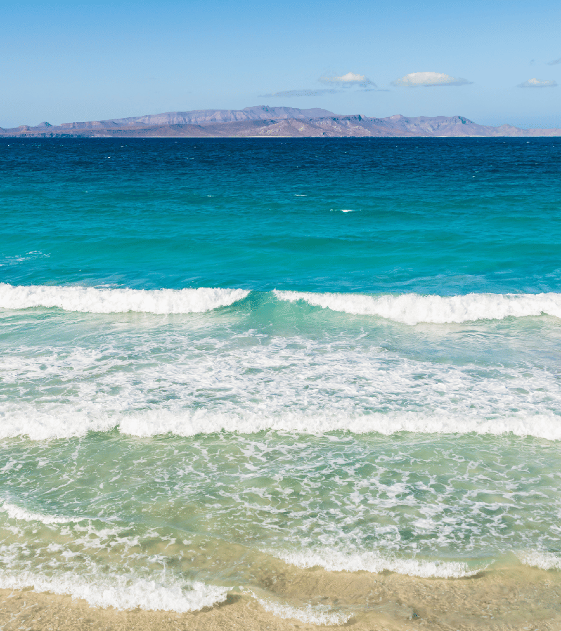 Waves gently breaking on a sandy beach with clear turquoise waters and a distant view of a rocky island under a bright blue sky.